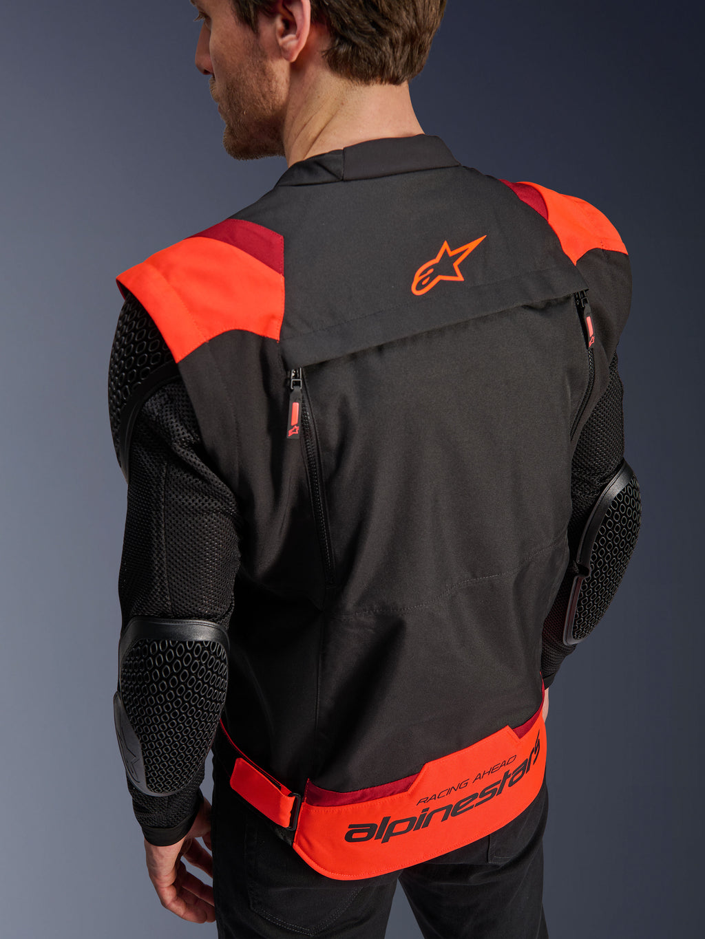 Kurtka Alpinestars T-Stunt Air, czarna z fluorescencyjną i ciemną czerwienią, widok z tyłu na modelu, siatkowe rękawy z ochraniaczami łokci o strukturze plastra miodu, logo gwiazdy Alpinestars na górze pleców, branding „RACING AHEAD” w dolnej części, modułowy sportowy krój.
