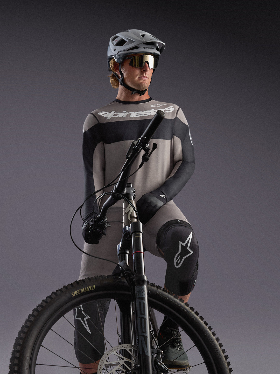 Szara koszulka Alpinestars A-Dura Race LS na rowerzyście MTB, czarne akcenty, profesjonalny sprzęt kolarski.