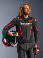 Skórzana kurtka Alpinestars GP Force V2, czarno-czerwona fluo, noszona przez modela trzymającego pasujący kask, białe logo Alpinestars i czerwone akcenty, w zestawieniu ze skórzanymi spodniami i rękawicami wyścigowymi, sportowa odzież motocyklowa na drogę i tor