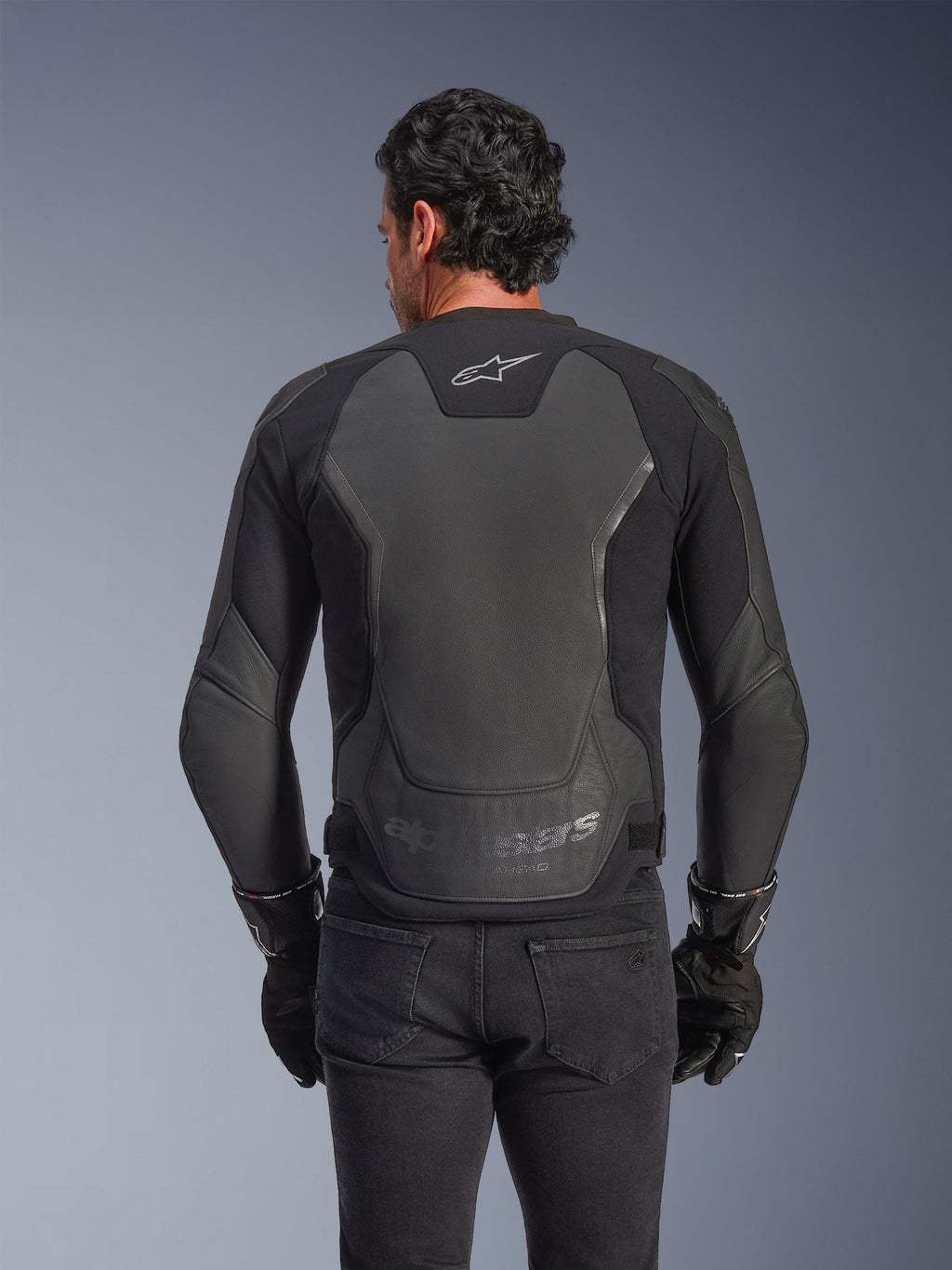 Alpinestars GP Force V2 Leather Jacket, czarna, widok z tyłu na mężczyznę w kurtce, czarnych rękawicach i dżinsach, duże skórzane panele ochronne na plecach, logo Alpinestars w górnej części pleców, wytłoczone oznaczenie marki w dolnej części talii, sportowa odzież motocyklowa