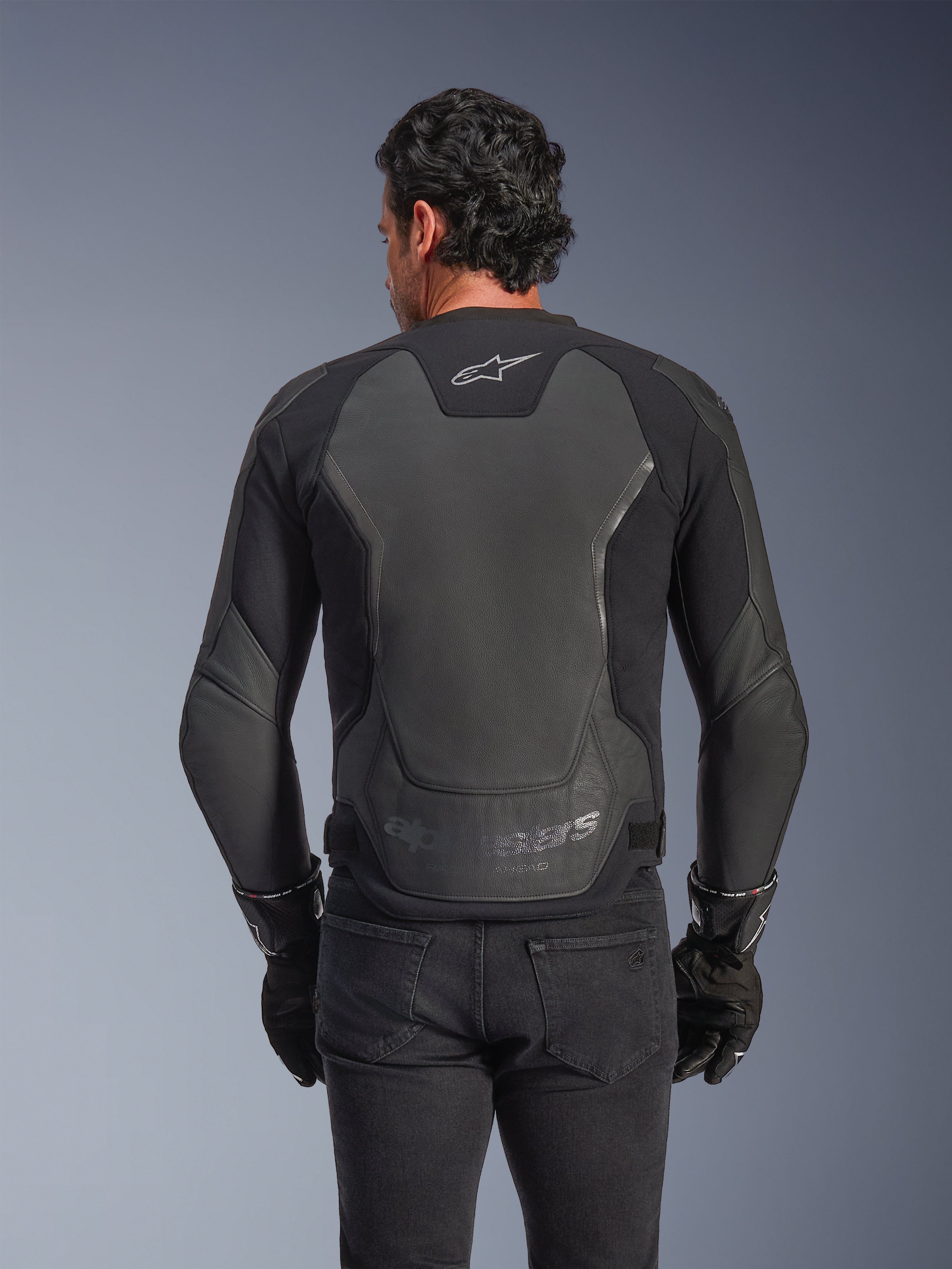 Alpinestars GP Force V2 Leather Jacket, czarna, widok z tyłu na mężczyznę w kurtce, czarnych rękawicach i dżinsach, duże skórzane panele ochronne na plecach, logo Alpinestars w górnej części pleców, wytłoczone oznaczenie marki w dolnej części talii, sportowa odzież motocyklowa
