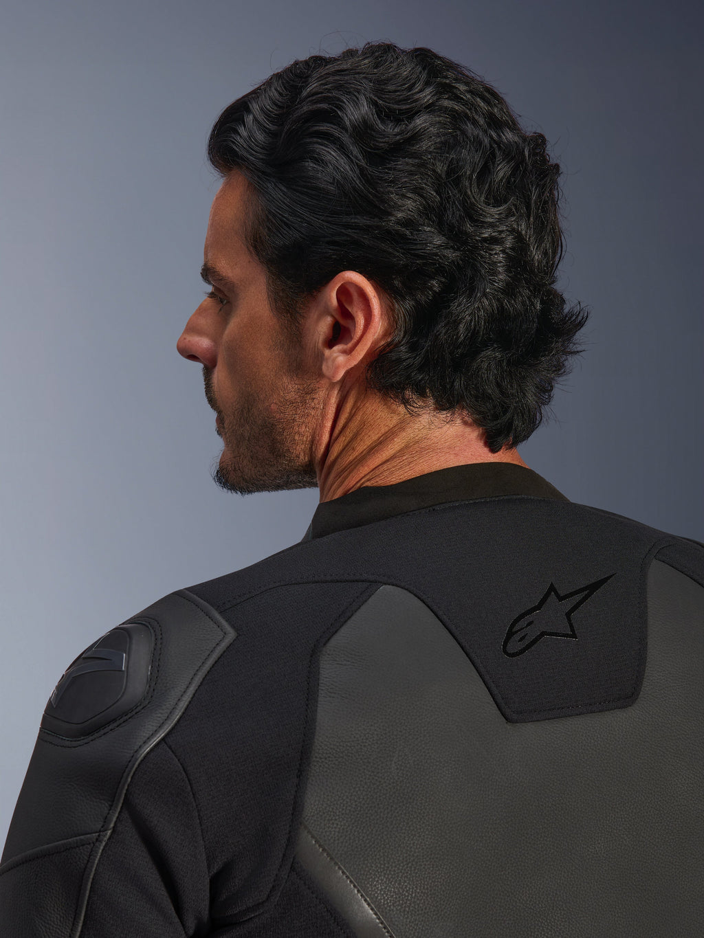 Alpinestars GP Force V2 Leather Jacket, czarna, zbliżenie z tyłu, z czarnym logo gwiazdy na górnej części pleców i ochraniaczem ramion GP Lite Evo DFS, wielopanelowa konstrukcja ze skóry i materiału tekstylnego, widok studyjny profilu motocyklisty i detali.