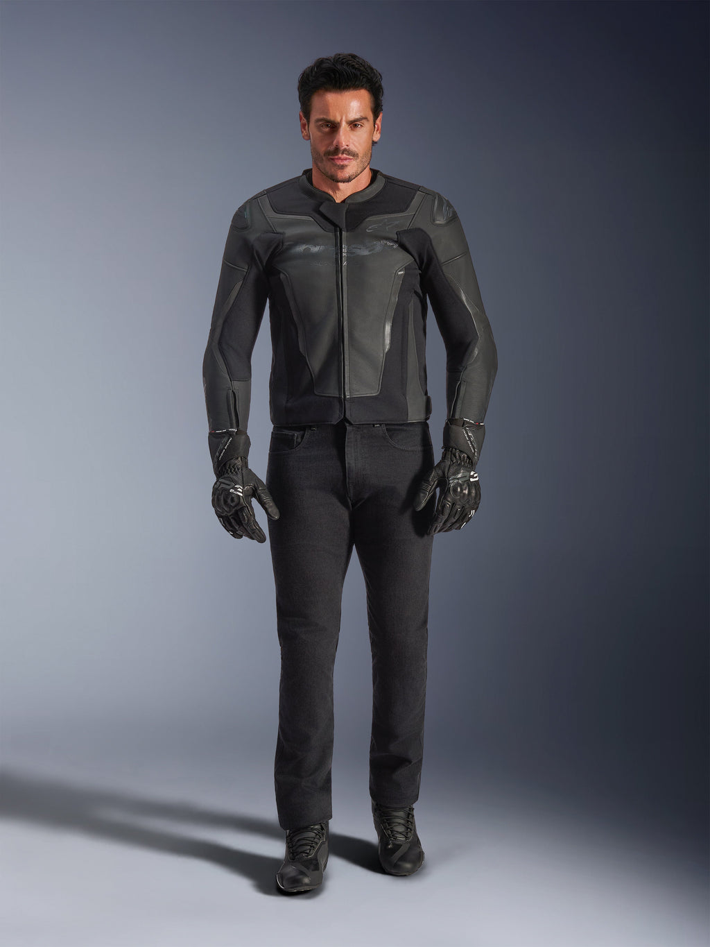Alpinestars GP Force V2 Leather Jacket w kolorze czarnym, noszona przez motocyklistę z czarnymi spodniami motocyklowymi, rękawicami i butami, smukła skórzana konstrukcja w sportowym stylu ze sliderami na ramionach i zintegrowaną ochroną