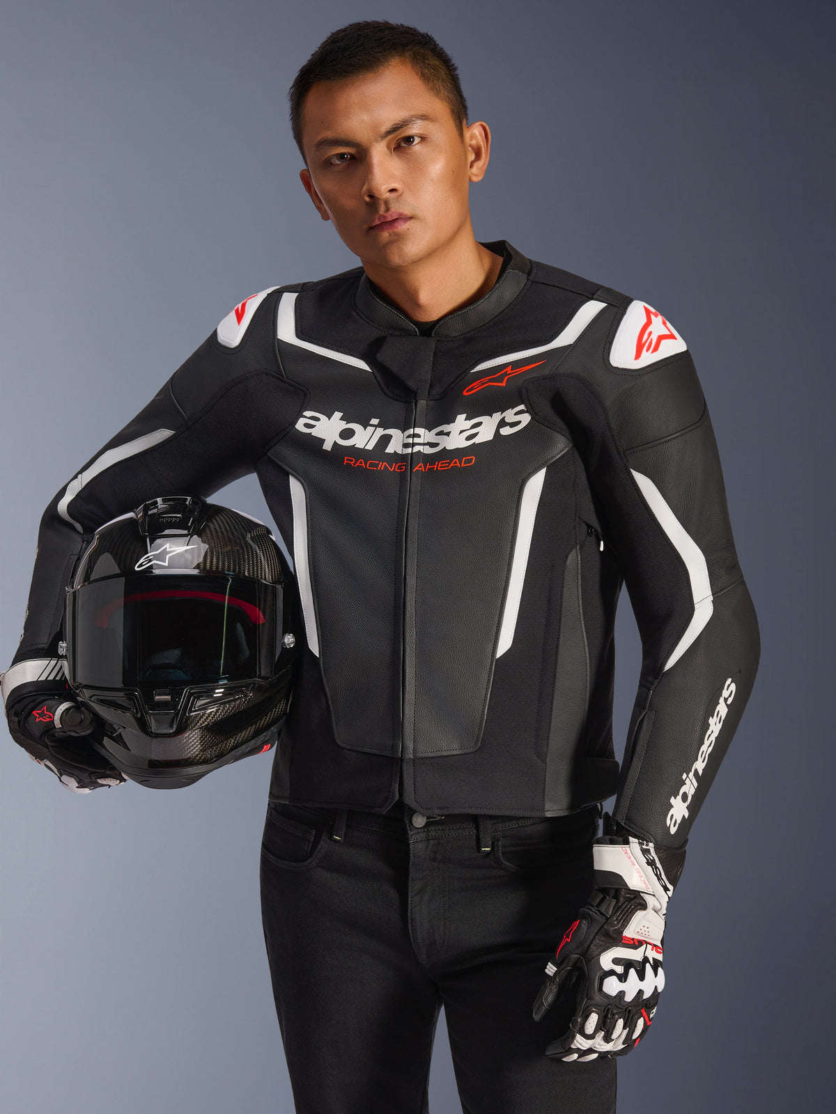 Alpinestars GP Force V2 Leather Jacket, skórzana kurtka motocyklowa, czarno-biała z czerwonymi akcentami, model trzymający kask motocyklowy z włókna węglowego i rękawice wyścigowe, białe kontrastowe panele i logo Alpinestars.