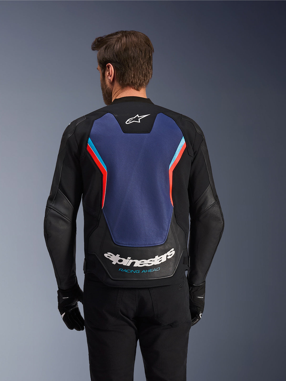 Widok z tyłu skórzanej kurtki Alpinestars GP Force V2 w kolorze czarnym, niebieskim i czerwonym fluo, z dużym niebieskim panelem na plecach, fluorescencyjnymi czerwonymi i jasnoniebieskimi akcentami, białym logo Alpinestars i napisem „Racing Ahead” w dolnej części pleców, prezentowana przez motocyklistę w czarnych rękawicach i spodniach.