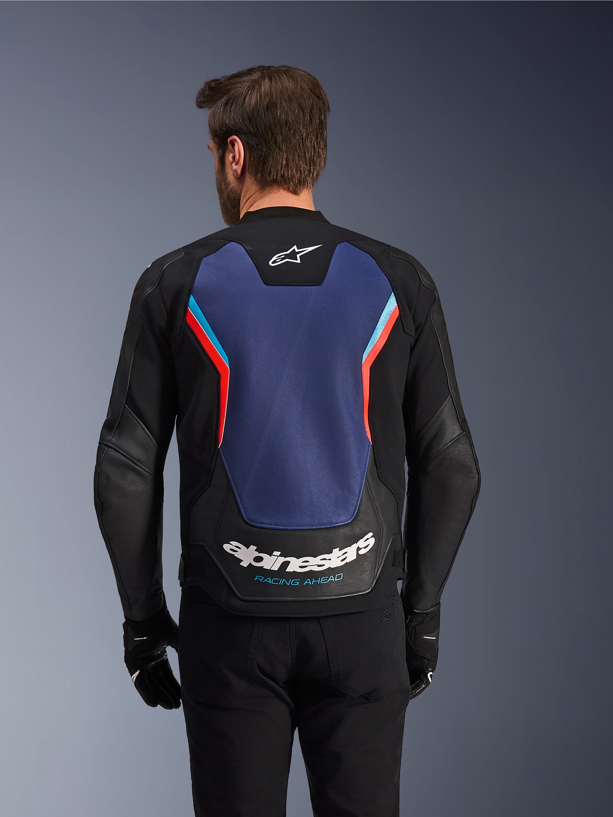 Rear view of the Alpinestars GP Force V2 Leather Jacket in Black, Blue, and Red Fluo, featuring a large blue back panel with fluorescent red and light blue accents, white Alpinestars logo and 'Racing Ahead' branding on the lower back, worn by a rider with black gloves and pants.