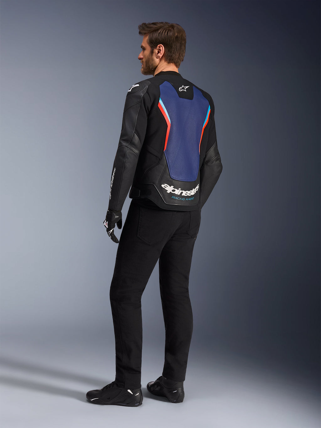 Widok z tyłu pod kątem trzech czwartych na modela ubranego w skórzaną kurtkę Alpinestars GP Force V2 w kolorystyce Black Blue Red Fluo, podkreślający niebieski panel na plecach z czerwonymi fluorescencyjnymi akcentami i białym logo, zestawiony z czarnymi spodniami i rękawicami motocyklowymi.