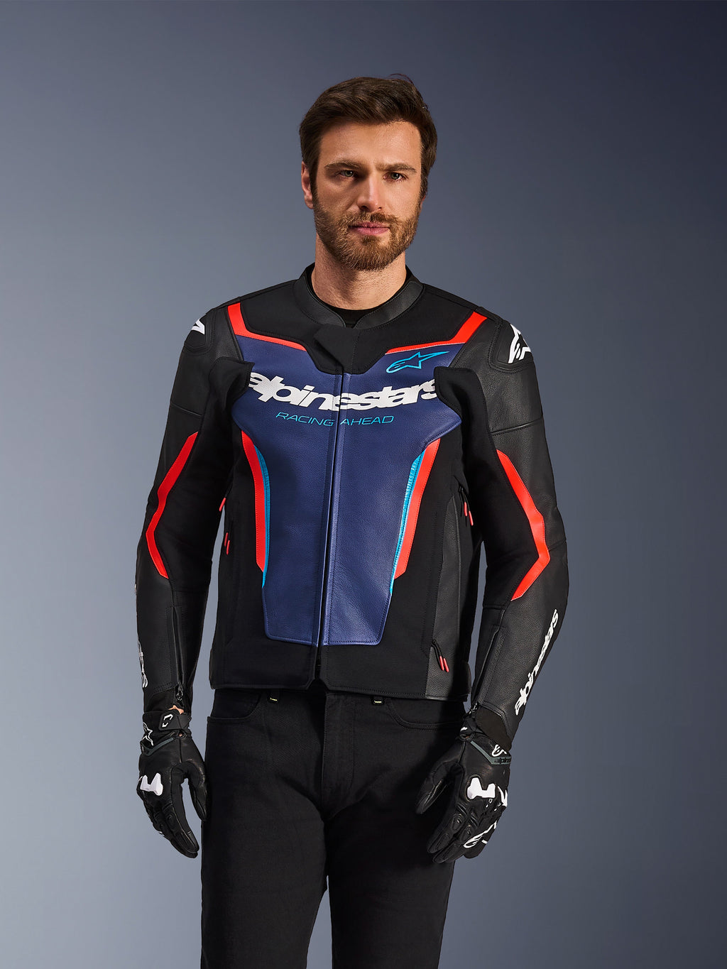 Kurtka skórzana Alpinestars GP Force V2, kolor czarny, niebieski i czerwony fluo, prezentowana przez modela w czarnych rękawicach i spodniach, niebieski panel na klatce piersiowej z napisem „Alpinestars Racing Ahead”, czerwone akcenty, zewnętrzne osłony ramion, sportowa odzież ochronna na motocykl