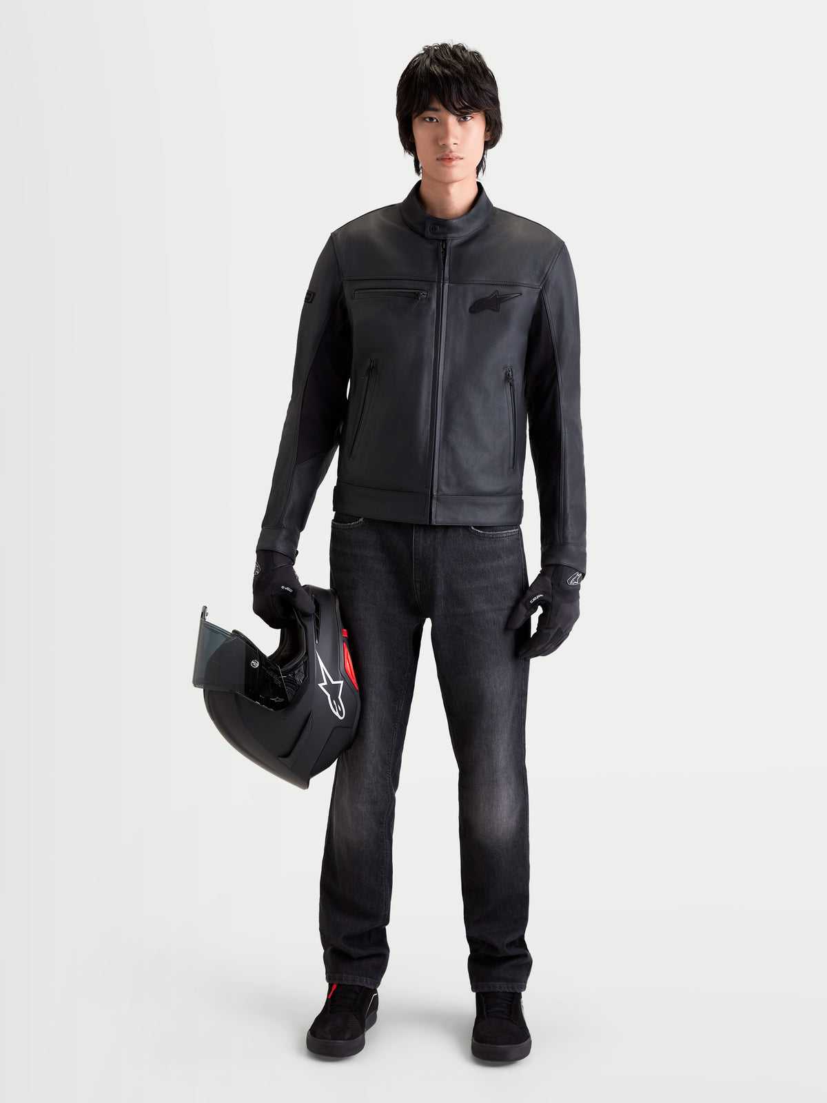 Alpinestars Maver X Kurtka skórzana, męska miejska motocyklowa kurtka skórzana w kolorze czarnym, widok z przodu na model w pasujących rękawicach, ciemnych jeansach i trzymający kask, z miękkiej skóry bydlęcej i z kołnierzem motocyklowym