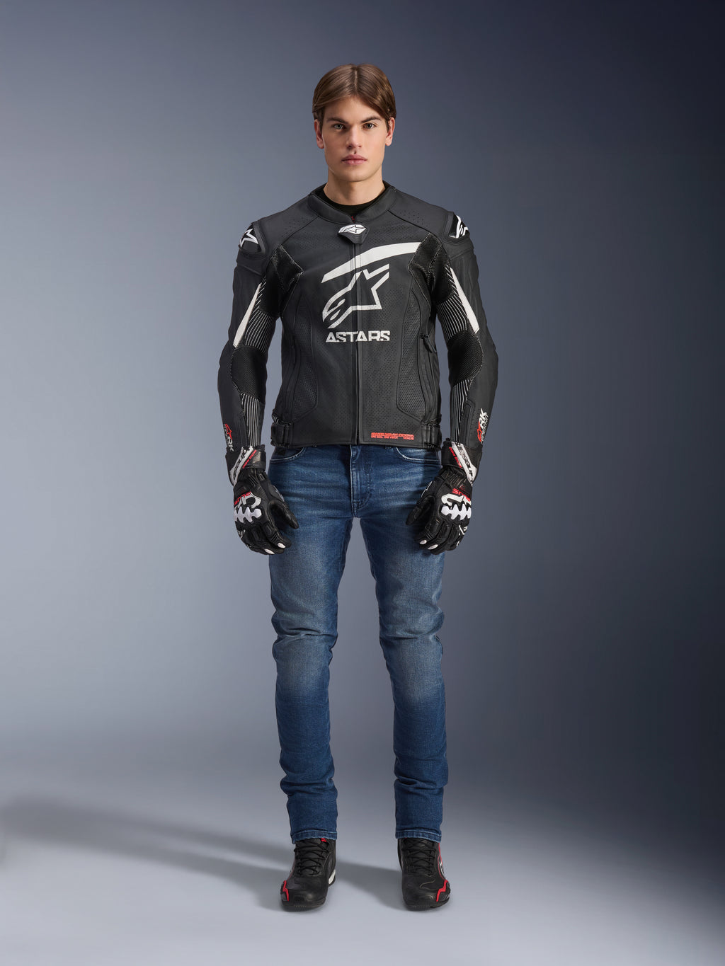 Męski model w skórzanej kurtce Alpinestars GP Plus R V4 Rideknit w kolorze czarno-białym, stylizowany z niebieskimi dżinsami motocyklowymi, czarnymi rękawicami i butami do jazdy, z dużym białym logo Astars na piersi, perforowanymi panelami skórzanymi i białymi pasami wyścigowymi na rękawach, widok całej sylwetki stojącej