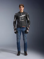 Męski model w skórzanej kurtce Alpinestars GP Plus R V4 Rideknit w kolorze czarno-białym, stylizowany z niebieskimi dżinsami motocyklowymi, czarnymi rękawicami i butami do jazdy, z dużym białym logo Astars na piersi, perforowanymi panelami skórzanymi i białymi pasami wyścigowymi na rękawach, widok całej sylwetki stojącej