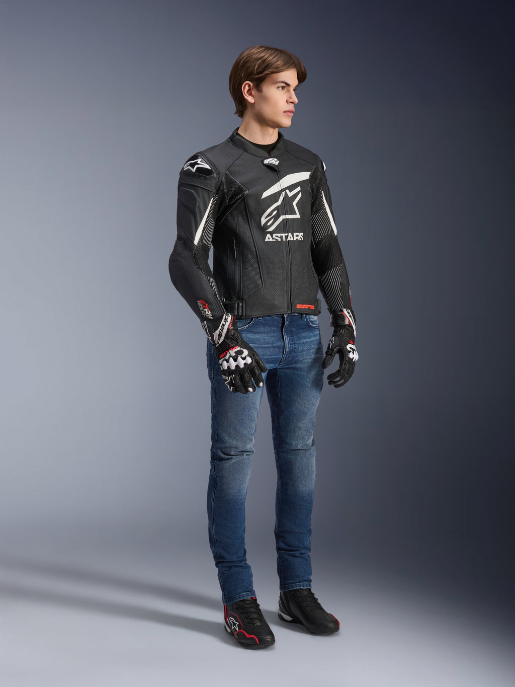 Kurtka skórzana Alpinestars GP Plus R V4 Rideknit, czarno-biała, motocyklowa kurtka skórzana noszona przez modela w niebieskich dżinsach i czarnych rękawicach, z wyraźnym białym logo Astars na klatce piersiowej, perforowanymi panelami i białymi akcentami na ramionach