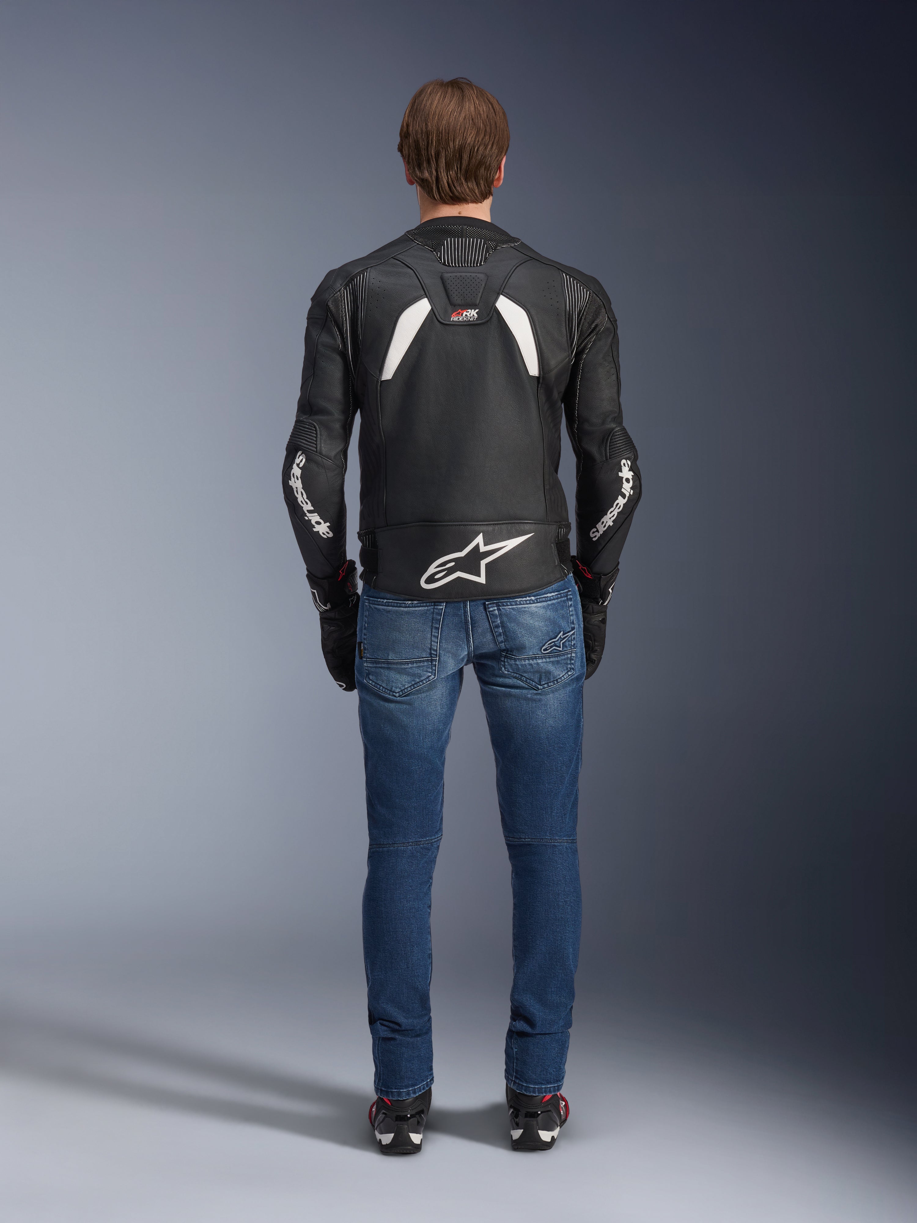Widok z tyłu skórzanej kurtki Alpinestars GP Plus R V4 Rideknit, czarno-biała, z dużym białym logo w dolnej części pleców i brandingiem na rękawach, noszona z niebieskimi jeansami motocyklowymi i czarnymi butami, podkreślająca techniczną konstrukcję i sportowy krój.