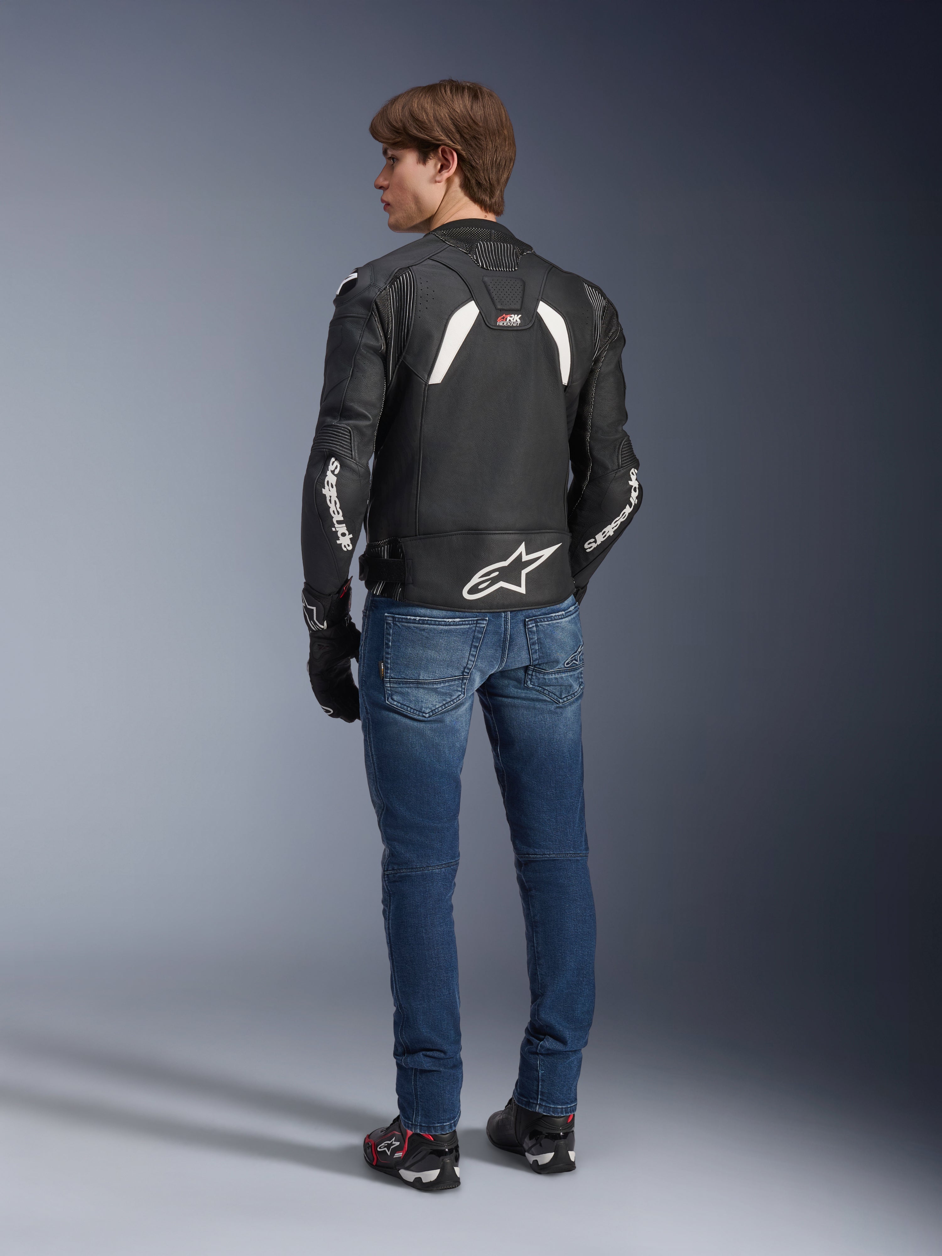 Alpinestars GP Plus R V4 Rideknit Leather Jacket, czarno-biała skórzana kurtka motocyklowa, widok z tyłu na modelu, biały branding Alpinestars na rękawach, duże logo gwiazdy w dolnej części pleców, niebieskie dżinsy, czarne buty motocyklowe, techniczna odzież motocyklowa
