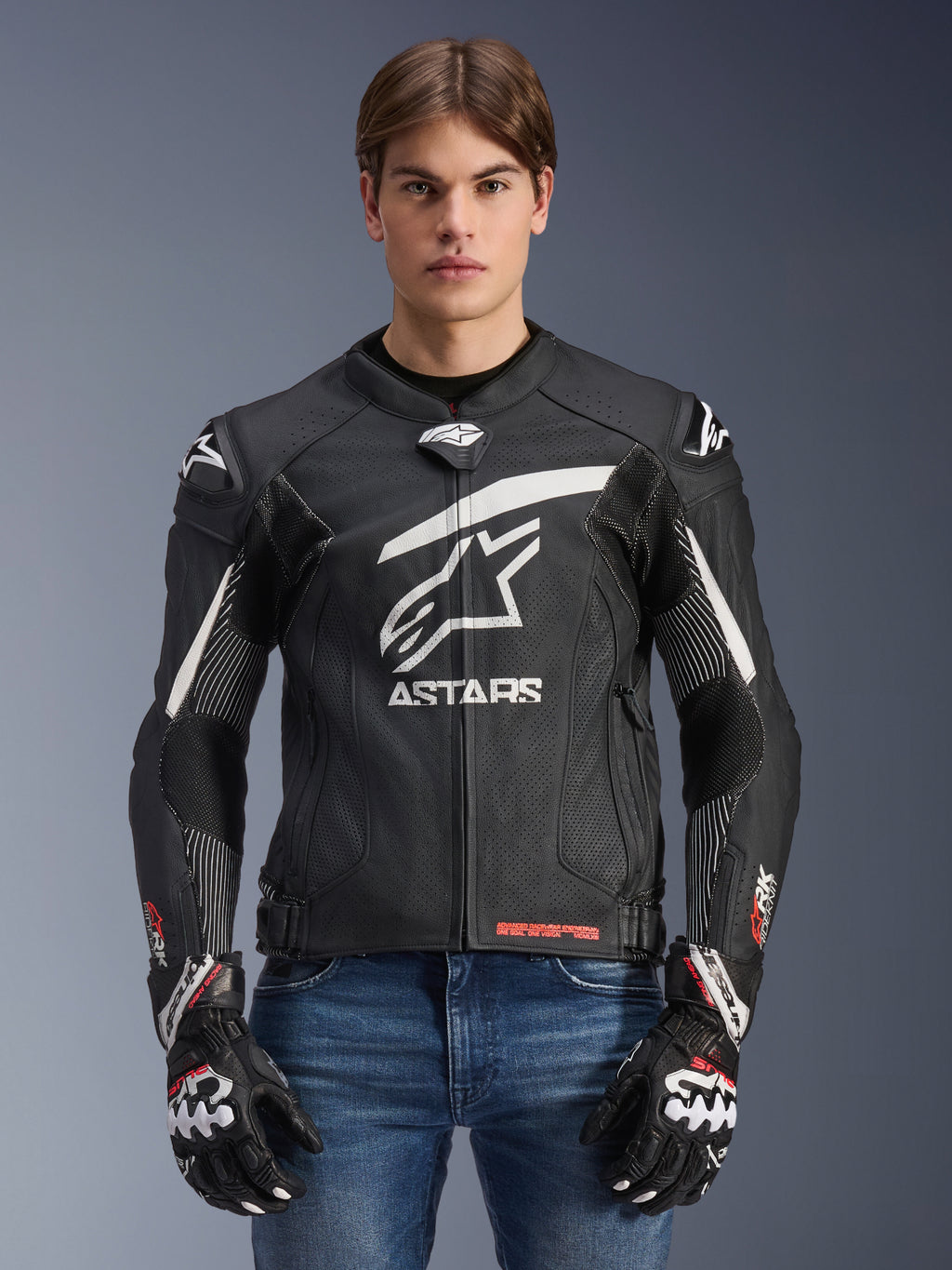 Skórzana kurtka Alpinestars GP Plus R V4 Rideknit, czarno-biała, motocyklowa kurtka wyścigowa noszona przez modela z dopasowanymi rękawicami i jeansami, z wyraźnym białym logo ASTARS na klatce piersiowej, perforowanymi panelami skórzanymi i ochronnymi sliderami na ramionach
