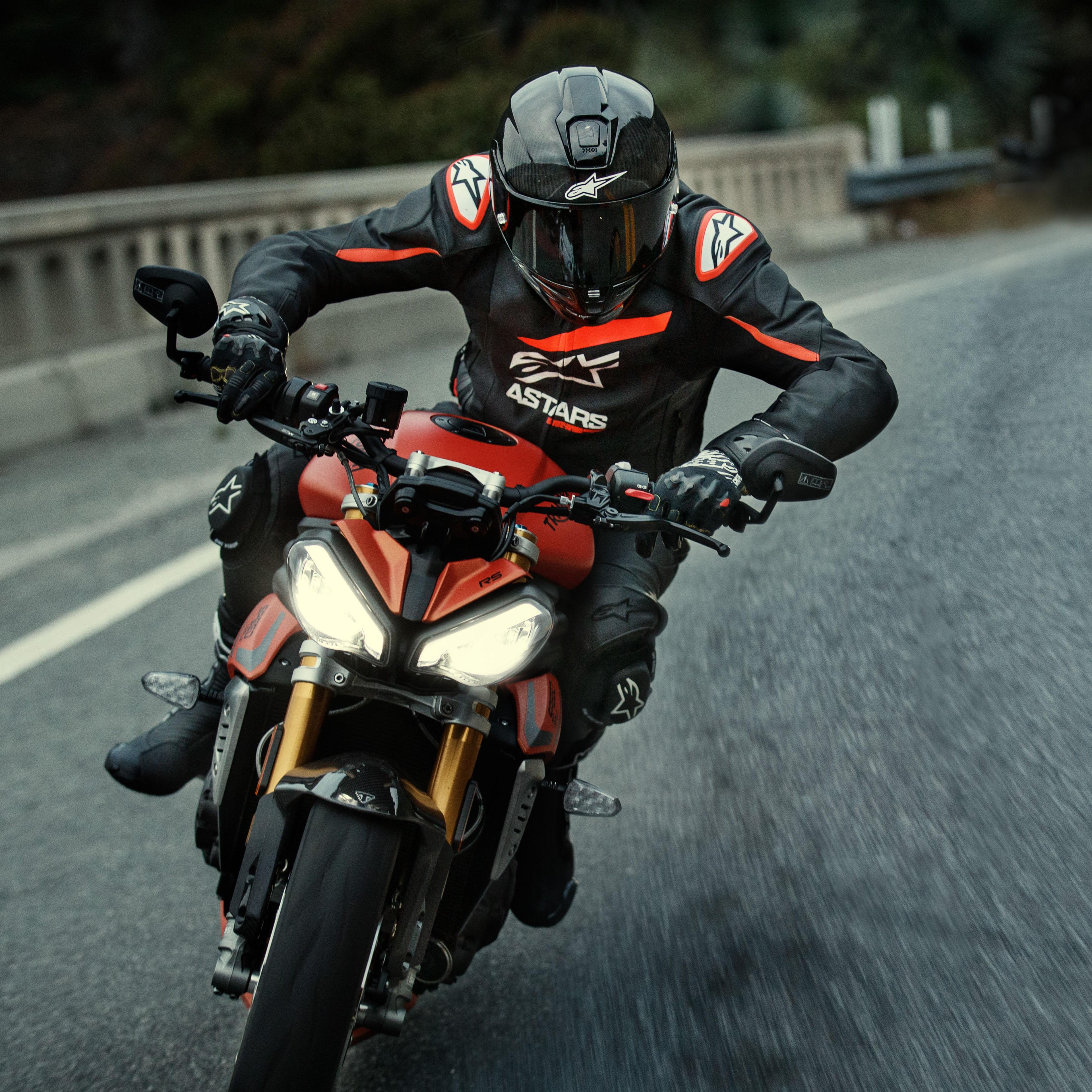 Motocyklista jadący na czerwonym motocyklu sportowym, skórzana kurtka Alpinestars GP Plus R V4 Rideknit, czarna z detalami w kolorze Red Fluo i białym, z białym logo Alpinestars i napisem „ASTARS”, w zestawie z czarnym kaskiem wyścigowym i rękawicami, wyczynowa odzież motocyklowa w akcji na drodze.