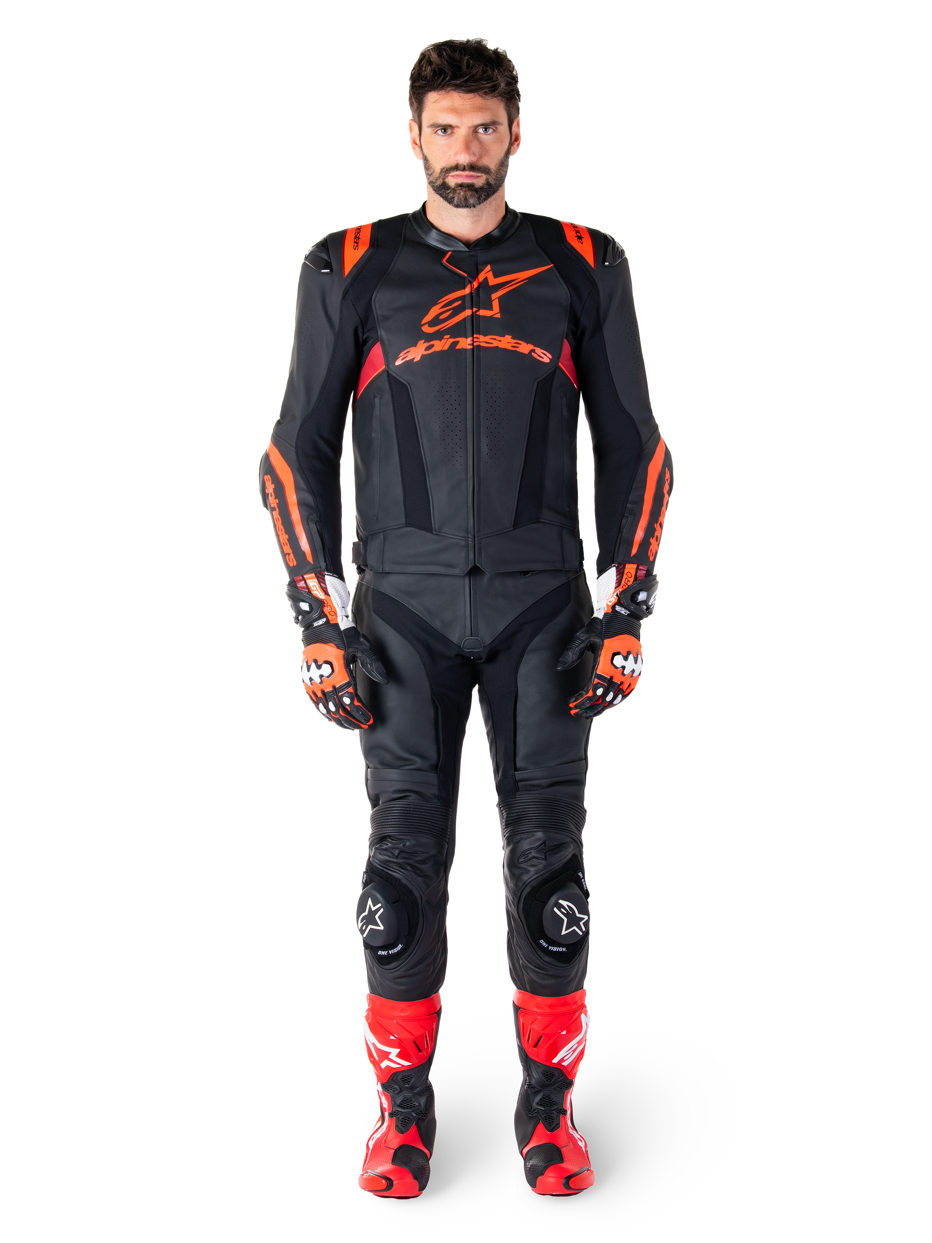 Męski model mający na sobie skórzaną kurtkę Alpinestars Missile V3 Ignition w kolorze Black Red Fluo Dark Red, kompletny motocyklowy strój wyścigowy, w tym pasujące skórzane spodnie, rękawice wyścigowe i buty, z wyraźnym brandingiem Alpinestars na klatce piersiowej, poza przodem, profesjonalny sportowy strój motocyklowy