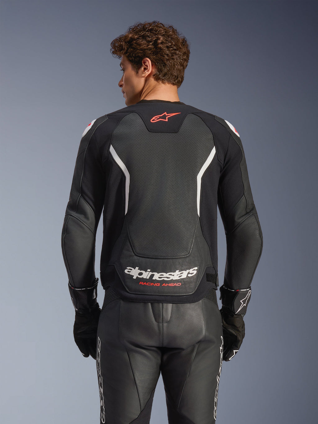 Alpinestars GP Force V2 Air Leather Jacket, skórzana kurtka motocyklowa, czarna z białymi i czerwonymi akcentami, widok od tyłu prezentujący perforowany skórzany panel na plecach, białe napisy Alpinestars i czerwone detale logo, noszona z pasującymi czarnymi skórzanymi spodniami wyścigowymi i rękawicami