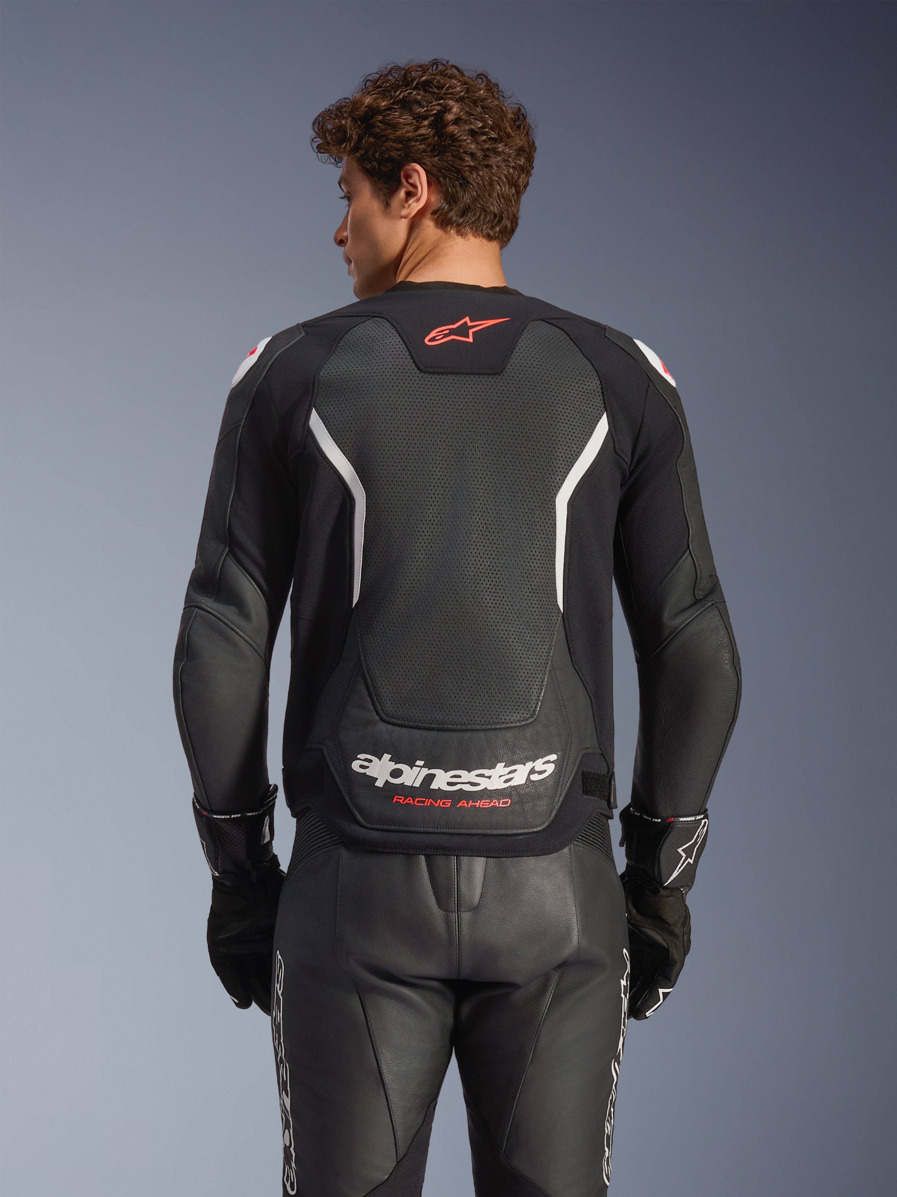 Alpinestars GP Force V2 Air Leather Jacket, skórzana kurtka motocyklowa, czarna z białymi i czerwonymi akcentami, widok od tyłu prezentujący perforowany skórzany panel na plecach, białe napisy Alpinestars i czerwone detale logo, noszona z pasującymi czarnymi skórzanymi spodniami wyścigowymi i rękawicami