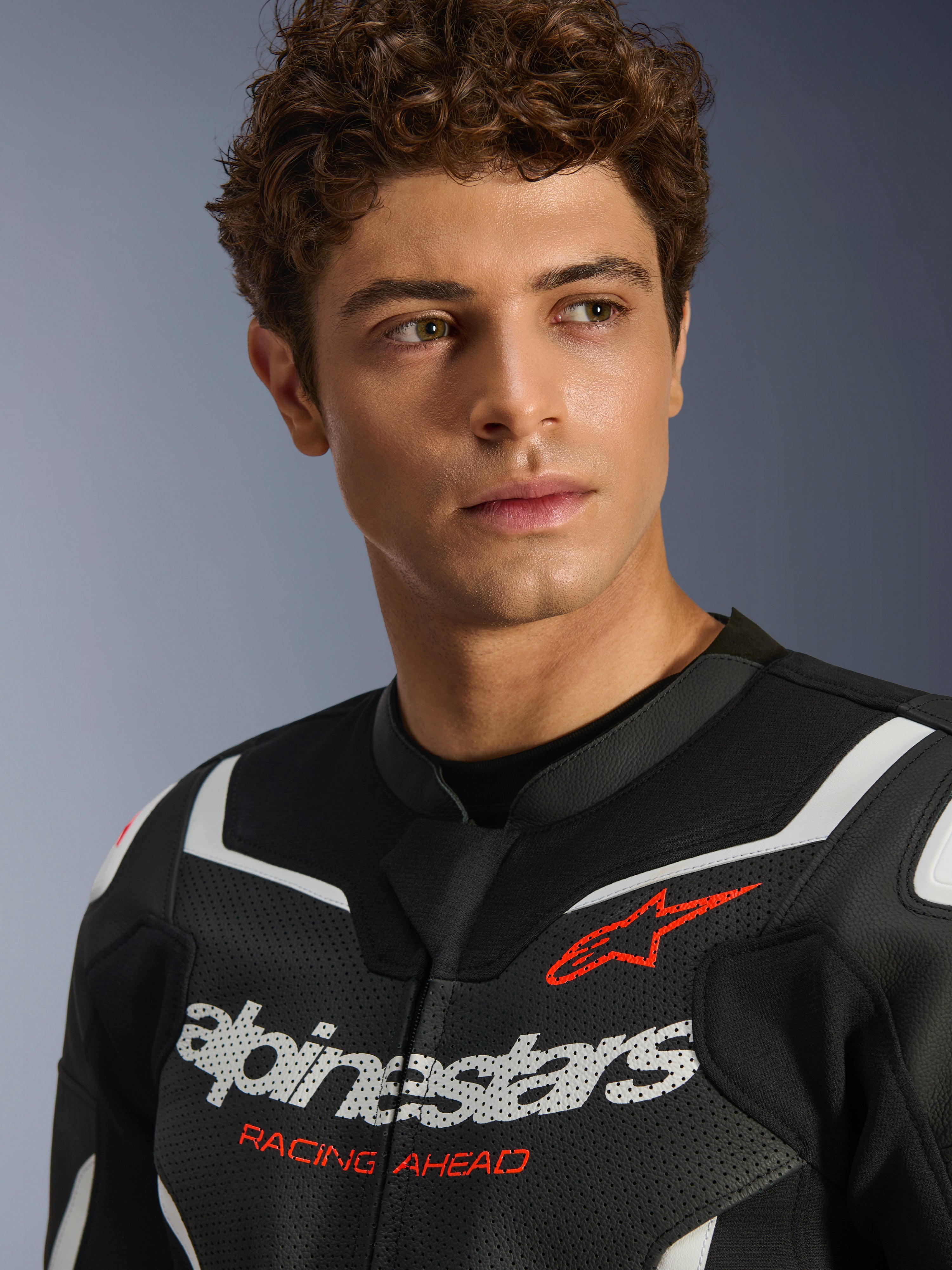 Portret męskiego modela w skórzanej kurtce Alpinestars GP Force V2 Air w kolorze czarno-białym, z perforowanej skóry, z wyraźnym białym logo i czerwonym napisem „RACING AHEAD” na klatce piersiowej, sportowa odzież motocyklowa