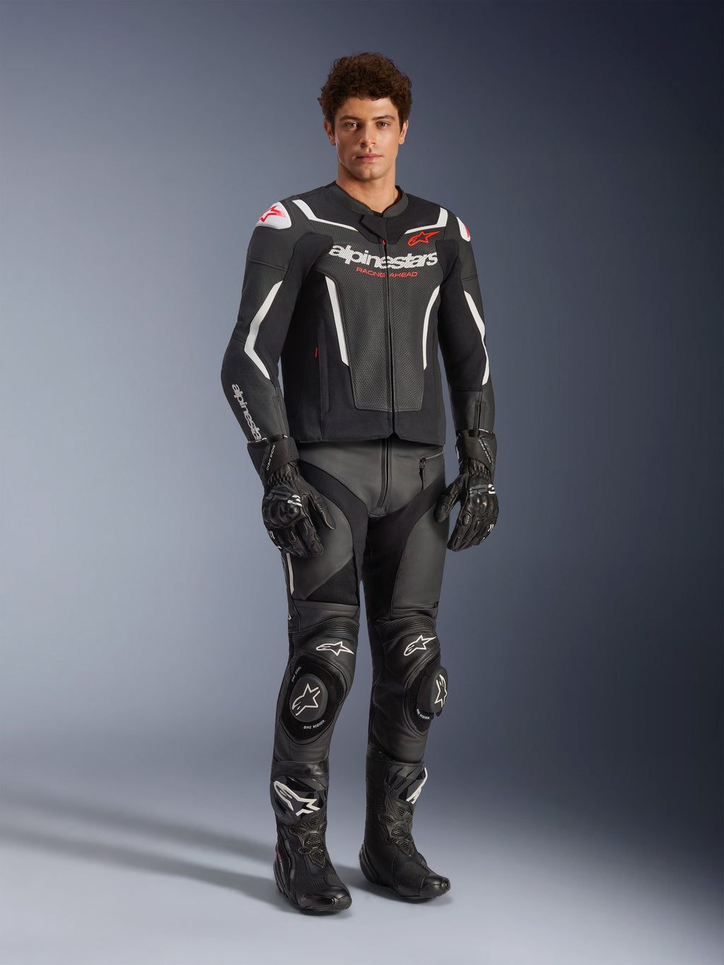 Kurtka skórzana Alpinestars GP Force V2 Air w kolorze czarno-białym, prezentowana przez motocyklistę z dopasowanymi czarnymi spodniami wyścigowymi, rękawicami i butami, wyposażona w perforowane panele skórzane, białe akcenty i czerwone logo Alpinestars na klatce piersiowej, sportowa odzież motocyklowa
