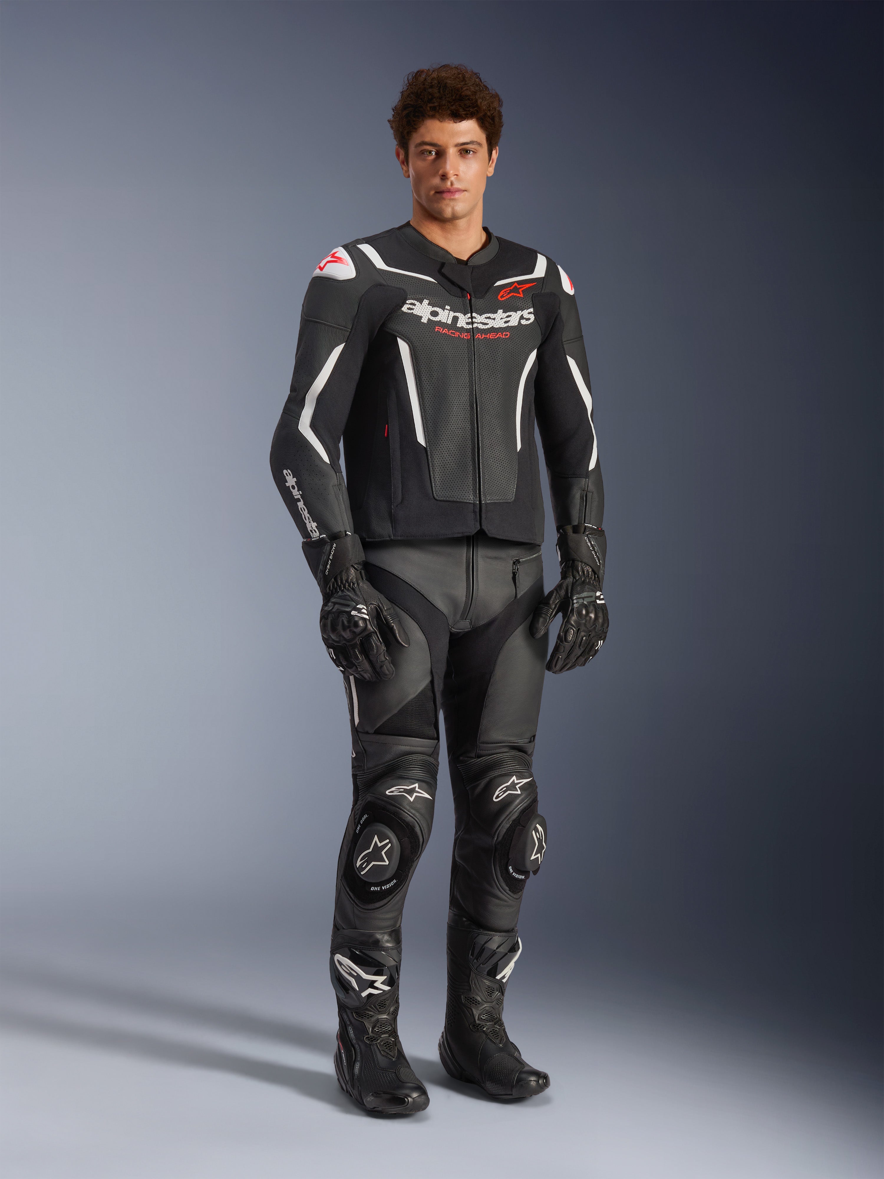 Kurtka skórzana Alpinestars GP Force V2 Air w kolorze czarno-białym, prezentowana przez motocyklistę z dopasowanymi czarnymi spodniami wyścigowymi, rękawicami i butami, wyposażona w perforowane panele skórzane, białe akcenty i czerwone logo Alpinestars na klatce piersiowej, sportowa odzież motocyklowa