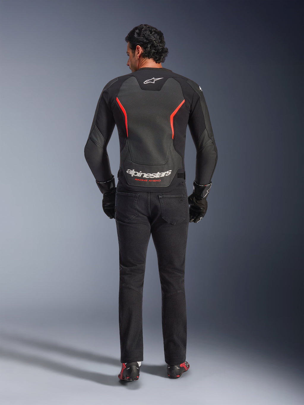 Skórzana kurtka Alpinestars GP Force V2 Air w kolorze czarno-czerwonym fluo, widok z tyłu na modelu, wyposażona w perforowane skórzane panele na plecach, czerwone akcenty i branding Alpinestars, w stylizacji z czarnymi jeansami motocyklowymi i rękawicami dla sportowego motocyklowego wyglądu.