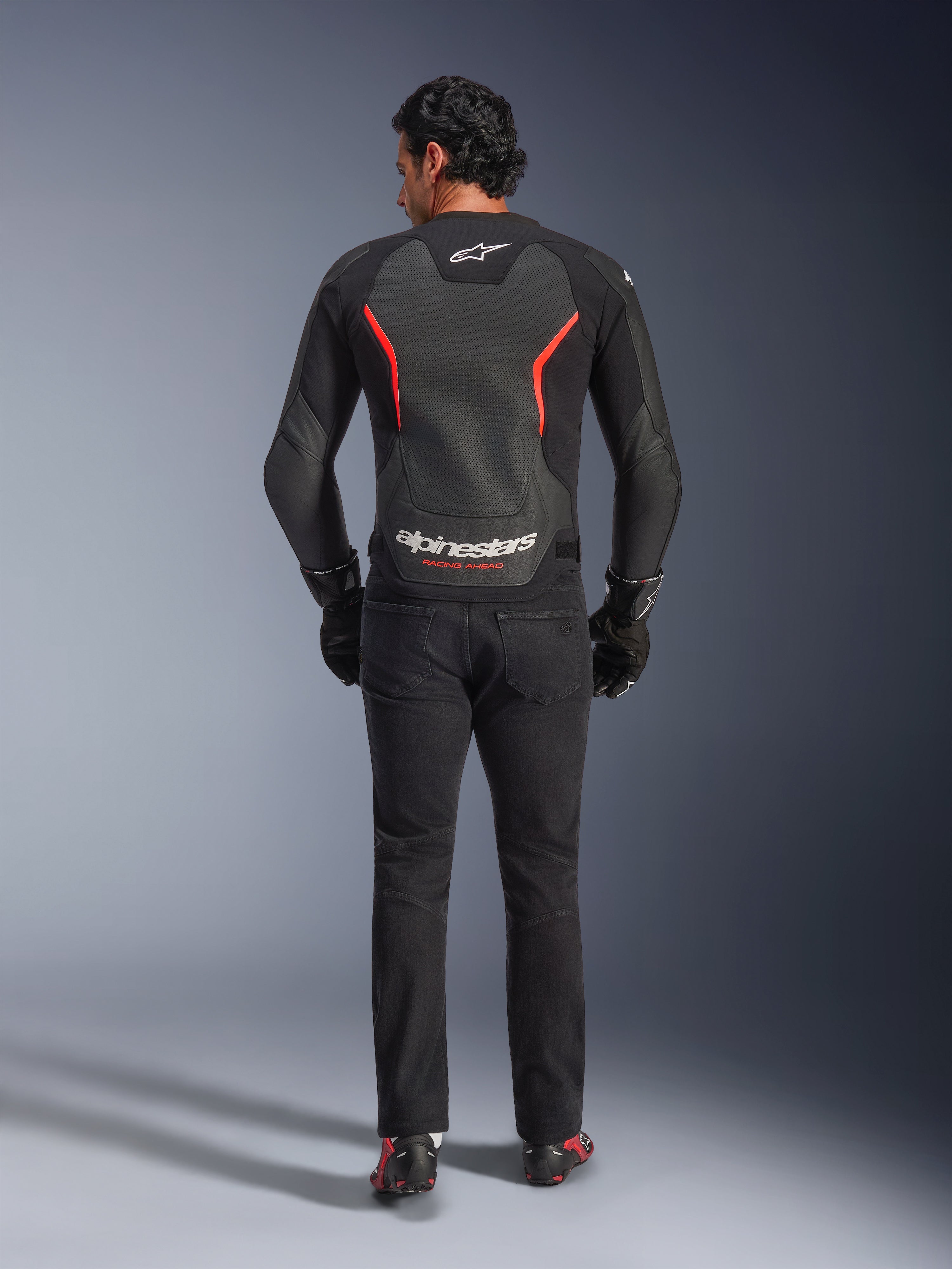 Skórzana kurtka Alpinestars GP Force V2 Air w kolorze czarno-czerwonym fluo, widok z tyłu na modelu, wyposażona w perforowane skórzane panele na plecach, czerwone akcenty i branding Alpinestars, w stylizacji z czarnymi jeansami motocyklowymi i rękawicami dla sportowego motocyklowego wyglądu.
