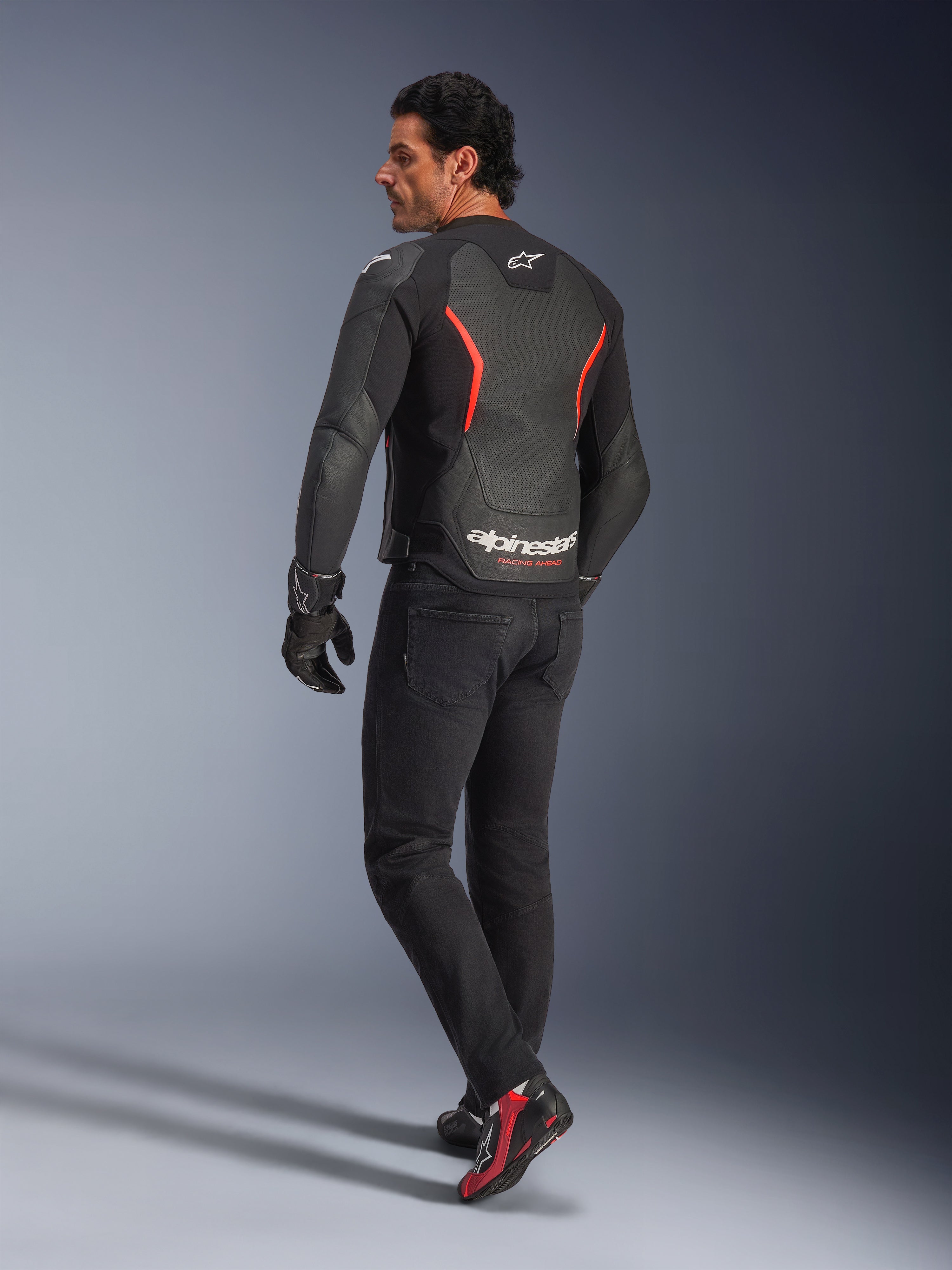 Męski model widoczny od tyłu w kurtce skórzanej Alpinestars GP Force V2 Air w kolorze Black Red Fluo, z licznymi perforacjami na plecach i wyraźnym białym brandingiem, zestawionej z czarnymi spodniami motocyklowymi, rękawicami i czarno-czerwonymi butami motocyklowymi, tło studyjne