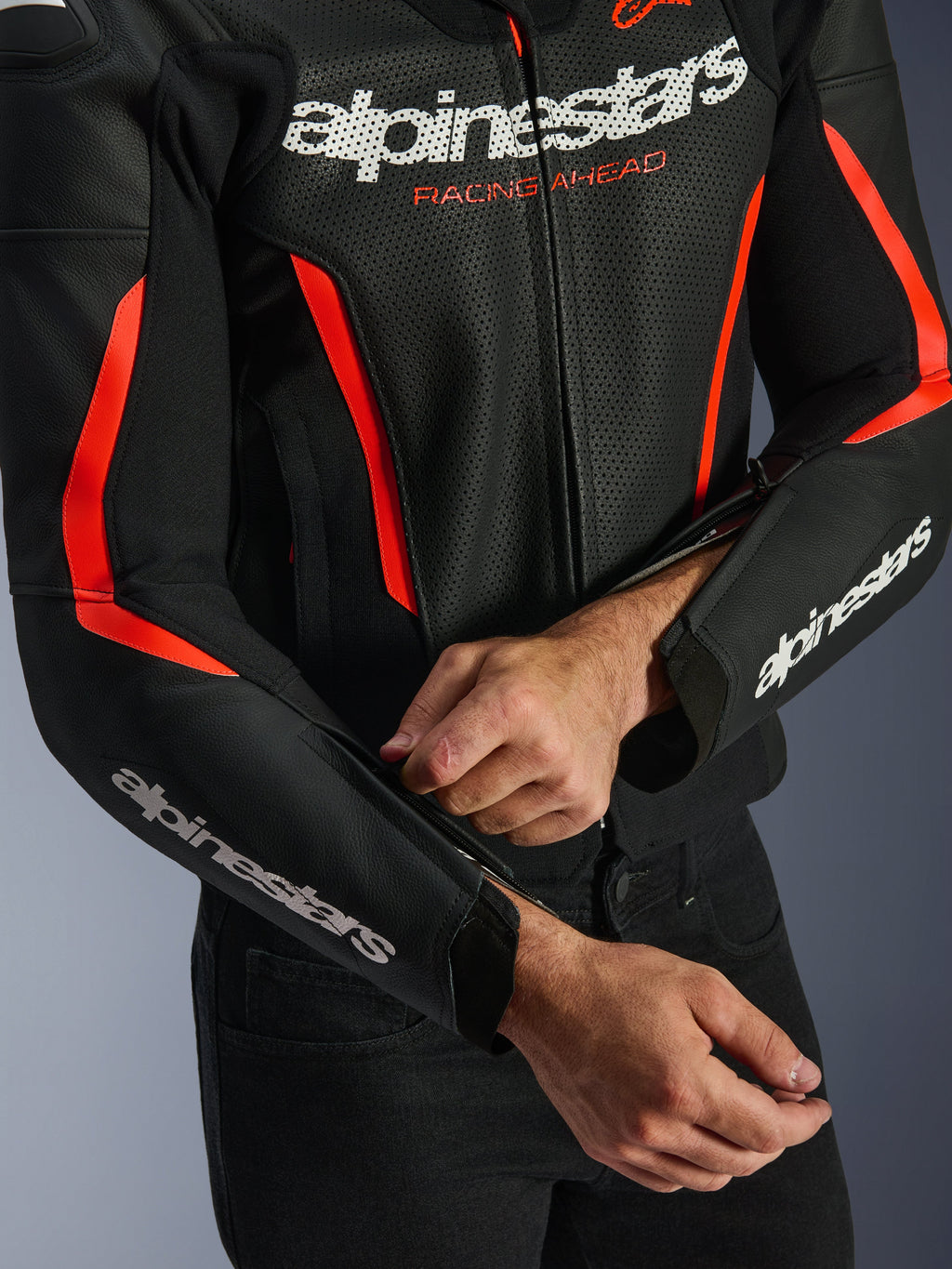 Skórzana kurtka Alpinestars GP Force V2 Air w kolorze czarnym i czerwonym fluo, zbliżenie na perforowaną konstrukcję skórzaną, białe logo na klatce piersiowej i rękawach, motocyklista regulujący zamek na przedramieniu dla lepszego dopasowania.