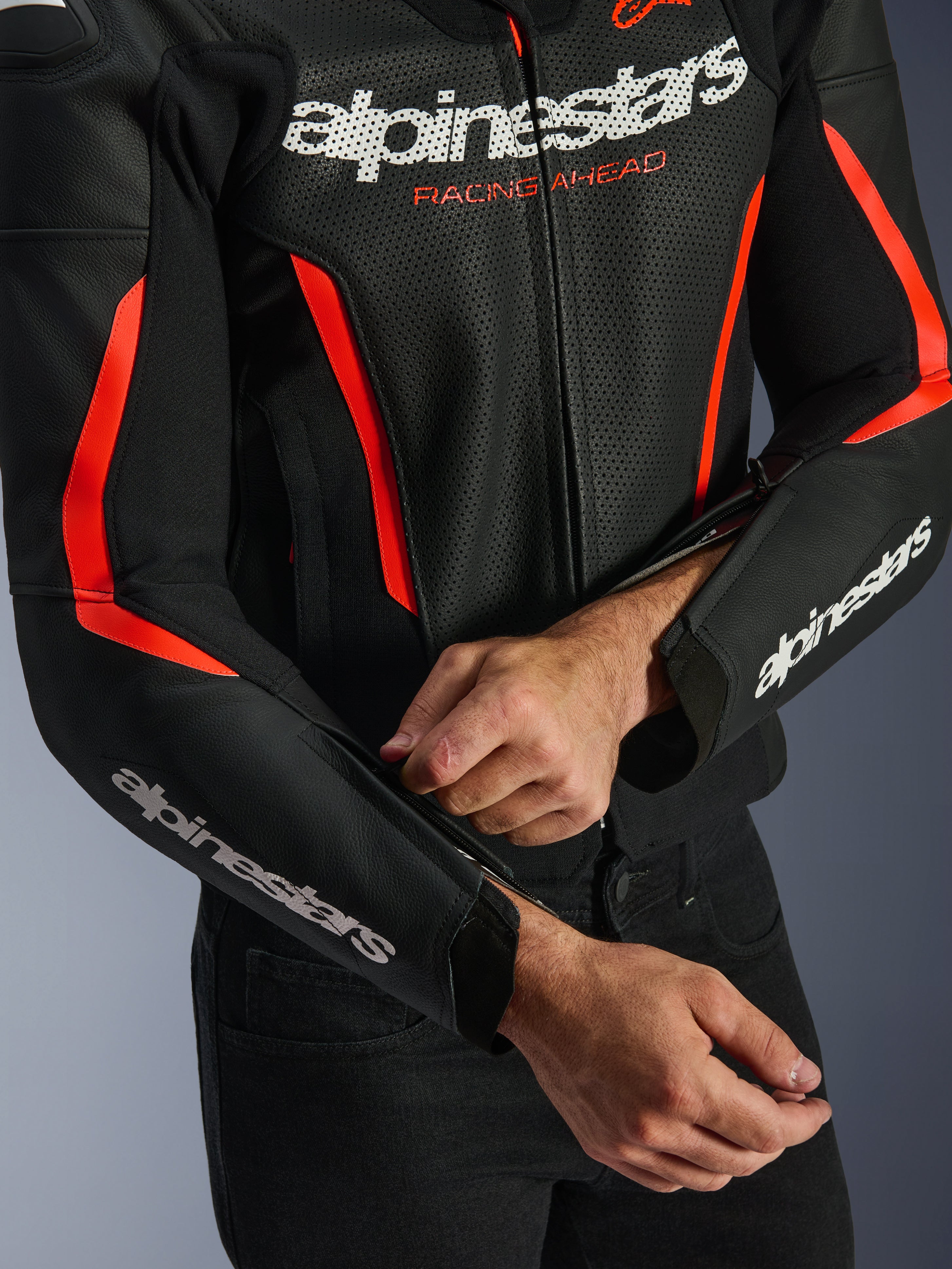 Skórzana kurtka Alpinestars GP Force V2 Air w kolorze czarnym i czerwonym fluo, zbliżenie na perforowaną konstrukcję skórzaną, białe logo na klatce piersiowej i rękawach, motocyklista regulujący zamek na przedramieniu dla lepszego dopasowania.