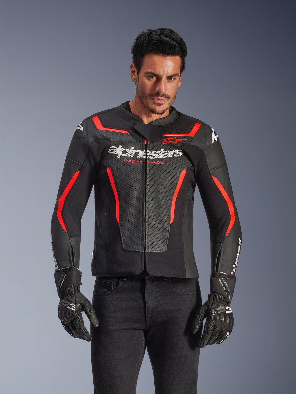 Skórzana kurtka motocyklowa Alpinestars GP Force V2 Air, kurtka wyścigowa, czarna i czerwona fluo, konstrukcja ze skóry premium z licznymi perforacjami zapewniającymi przepływ powietrza, noszona przez modela w czarnych rękawicach i spodniach, z wyraźnym brandingiem i napisem „Racing Ahead” na klatce piersiowej