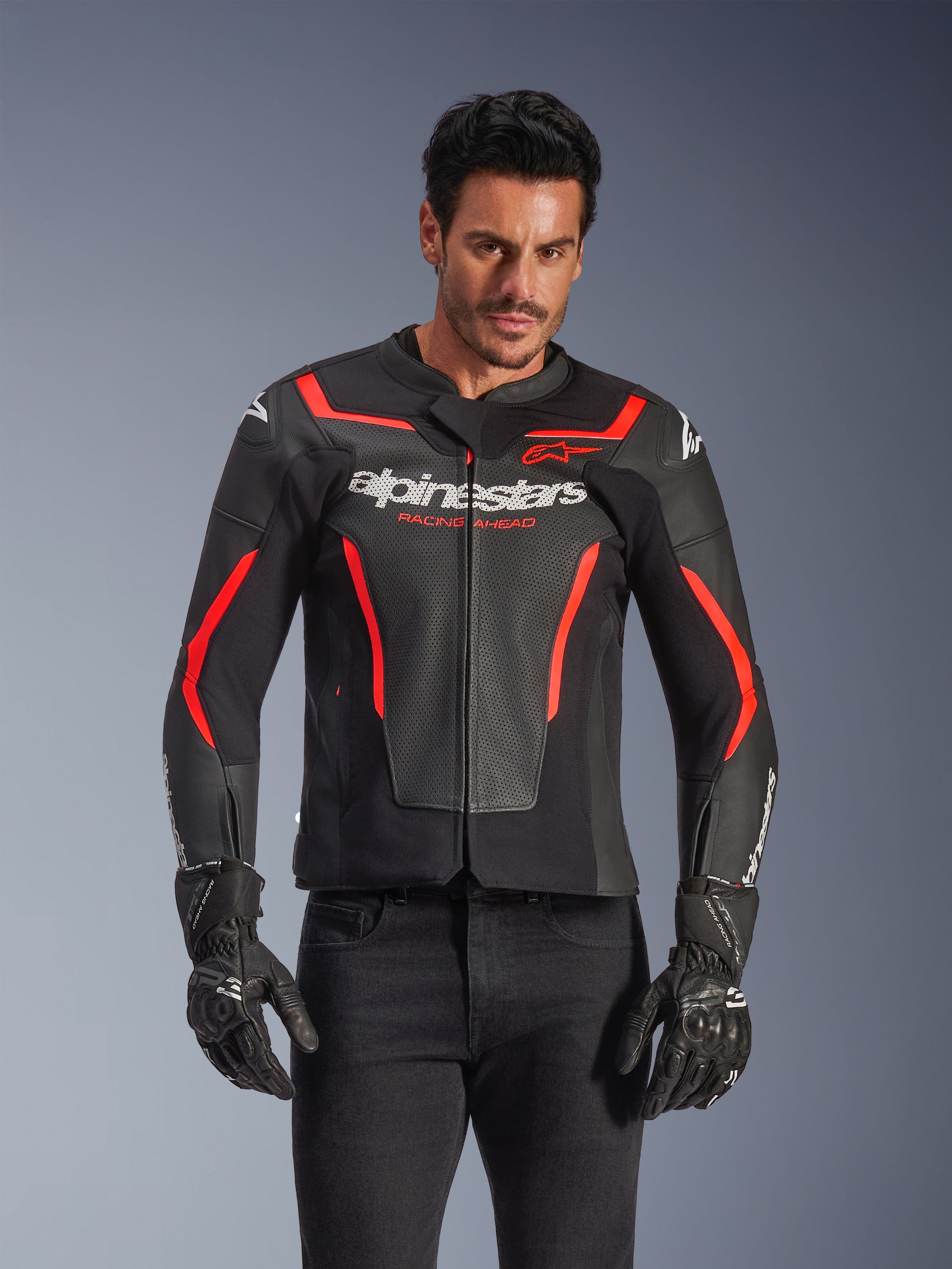 Skórzana kurtka motocyklowa Alpinestars GP Force V2 Air, kurtka wyścigowa, czarna i czerwona fluo, konstrukcja ze skóry premium z licznymi perforacjami zapewniającymi przepływ powietrza, noszona przez modela w czarnych rękawicach i spodniach, z wyraźnym brandingiem i napisem „Racing Ahead” na klatce piersiowej