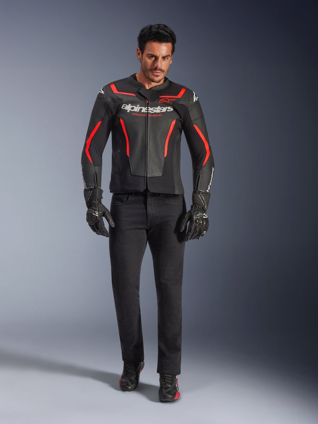 Motocyklista w skórzanej kurtce Alpinestars GP Force V2 Air, czarno-czerwona Fluo, zestawiona z czarnymi jeansami, rękawicami i butami, perforowana skórzana konstrukcja, białe logo i sportowe czerwone akcenty