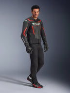 Model w kurtce skórzanej Alpinestars GP Force V2 Air w kolorze czarnym i czerwonym fluo, zestawionej z czarnymi spodniami motocyklowymi, rękawicami i pasującymi sneakersami, z perforowanymi panelami skórzanymi i wyraźnym białym logo Alpinestars, sportowa odzież motocyklowa