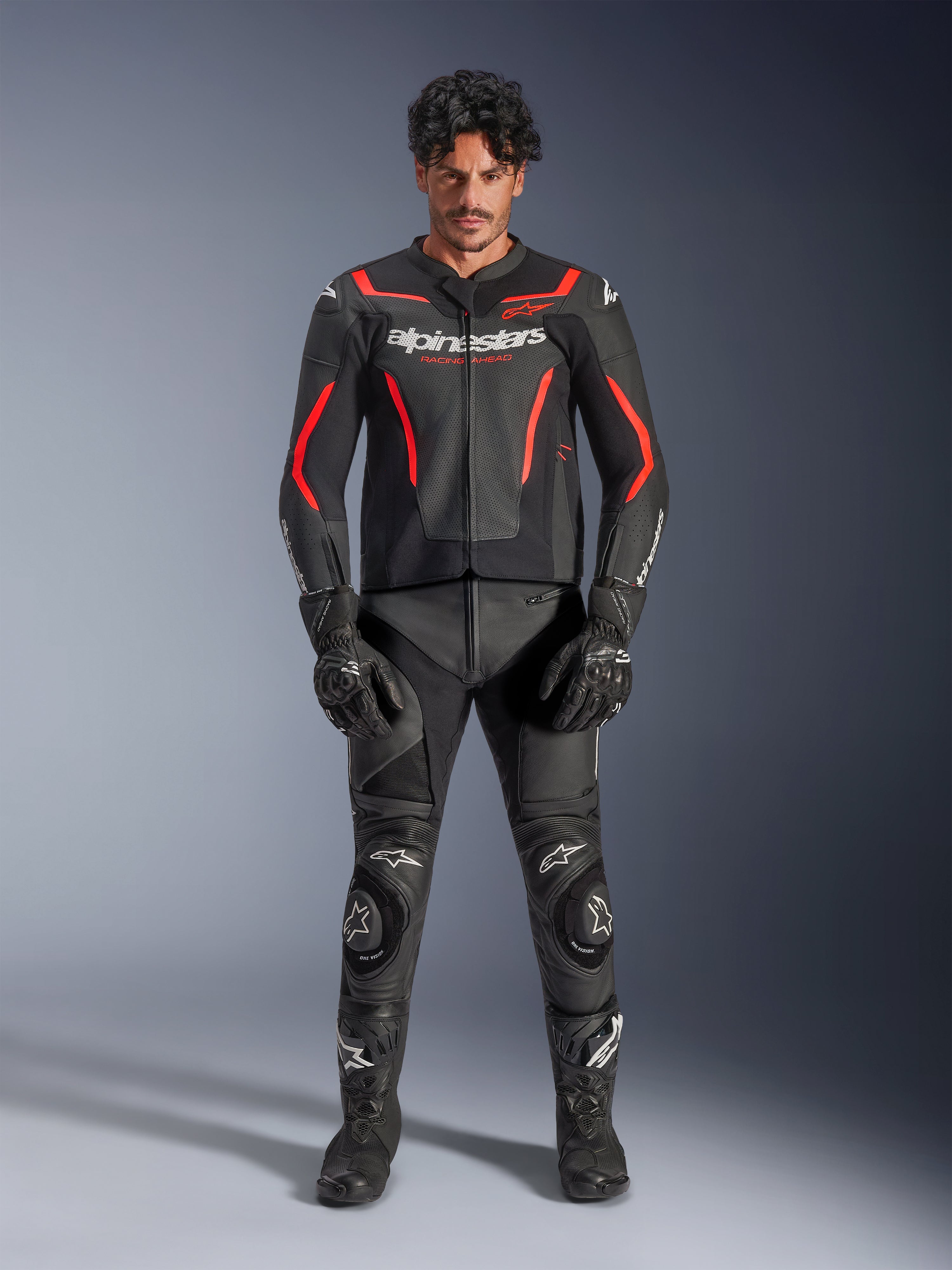 Męski model w skórzanej kurtce Alpinestars GP Force V2 Air w kolorze czarno-czerwonym (Black Red Fluo), z perforowanymi panelami na klatce piersiowej i białym logo, w zestawie z czarnymi skórzanymi spodniami motocyklowymi, czarnymi rękawicami i czarnymi butami, tworzący kompletny wyścigowy look.