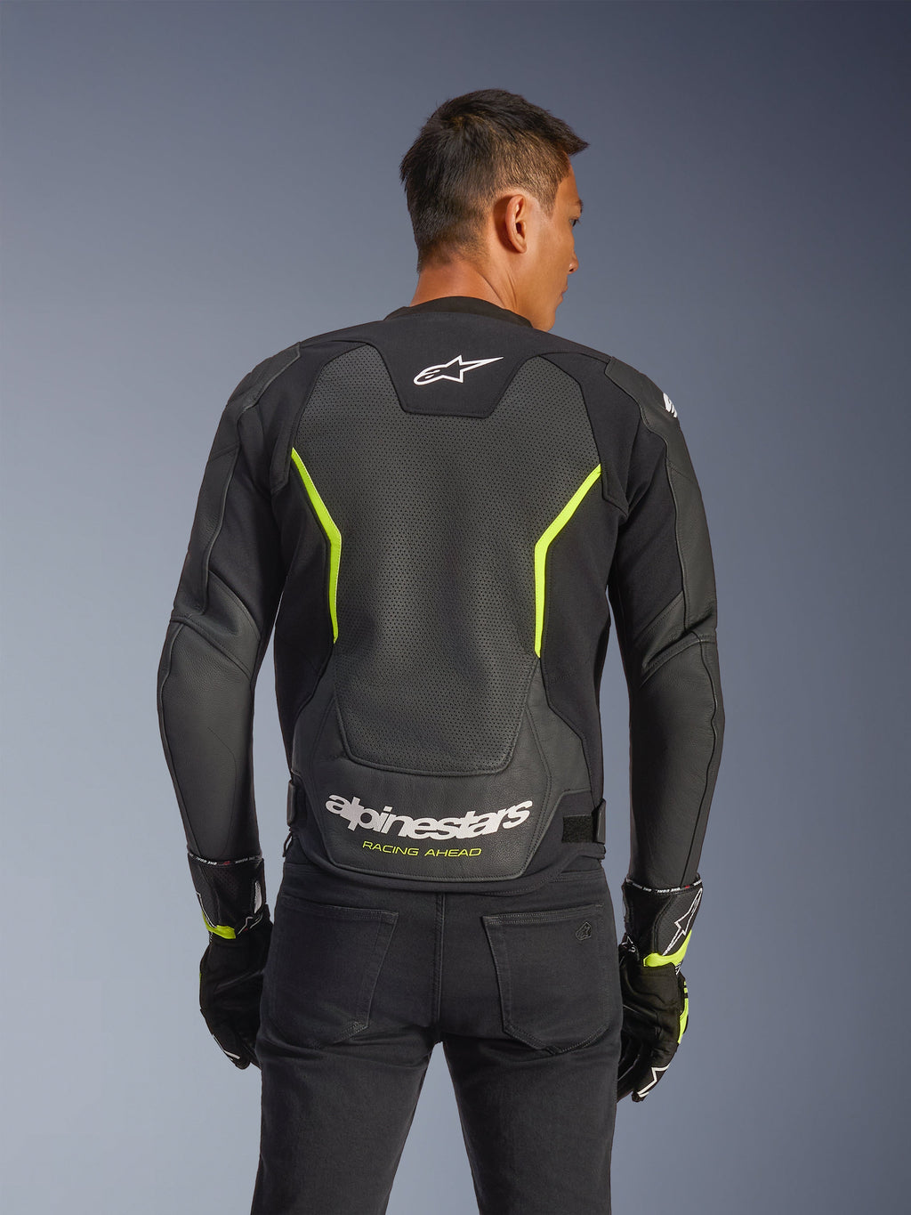 Alpinestars GP Force V2 Air Leather Jacket, czarna i żółta fluo, widok z tyłu na motocyklistę prezentujący rozległe perforowane panele skórzane zapewniające przepływ powietrza, białe logo Alpinestars i żółty napis „Racing Ahead” na dole pleców, sportowa odzież motocyklowa.