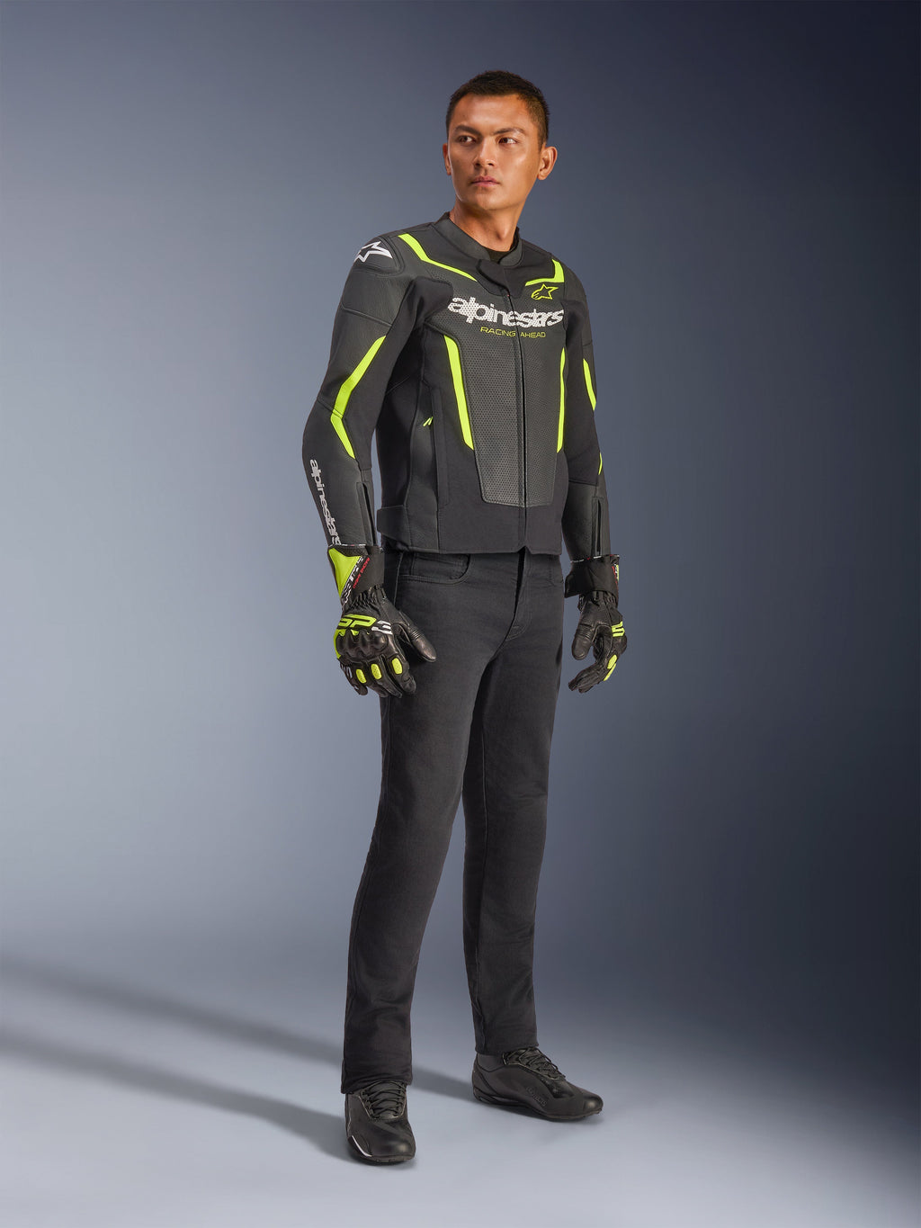 Męski model w skórzanej kurtce Alpinestars GP Force V2 Air w kolorze czarnym i żółtym fluo, zestawionej z czarnymi spodniami motocyklowymi, dopasowanymi rękawicami i czarnymi butami, ujęcie całej sylwetki w studio przedstawiające sportowy strój motocyklowy z perforowanej skóry i wyraźnym brandingiem