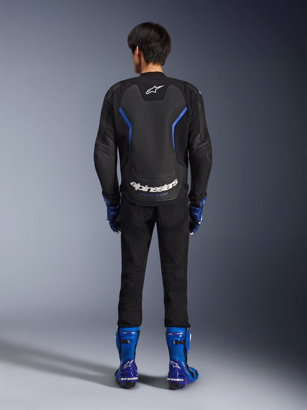 Skórzana kurtka Alpinestars GP Force V2 Airflow, Black Metallic Blue, widok z tyłu ukazujący perforowane panele skórzane i białe logotypy, noszona z niebieskimi butami wyścigowymi i czarnymi spodniami, sportowy strój motocyklowy