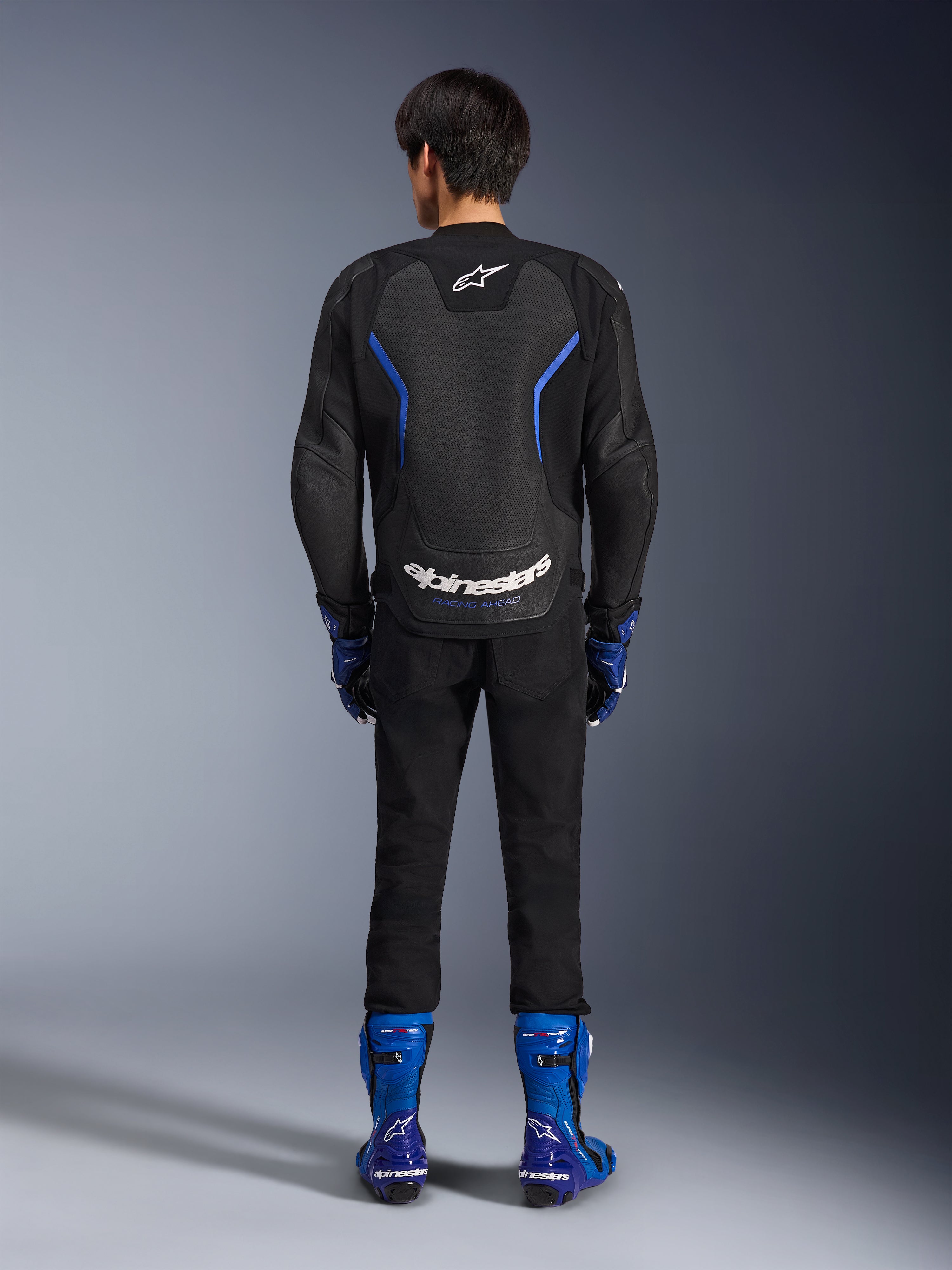 Skórzana kurtka Alpinestars GP Force V2 Airflow, Black Metallic Blue, widok z tyłu ukazujący perforowane panele skórzane i białe logotypy, noszona z niebieskimi butami wyścigowymi i czarnymi spodniami, sportowy strój motocyklowy