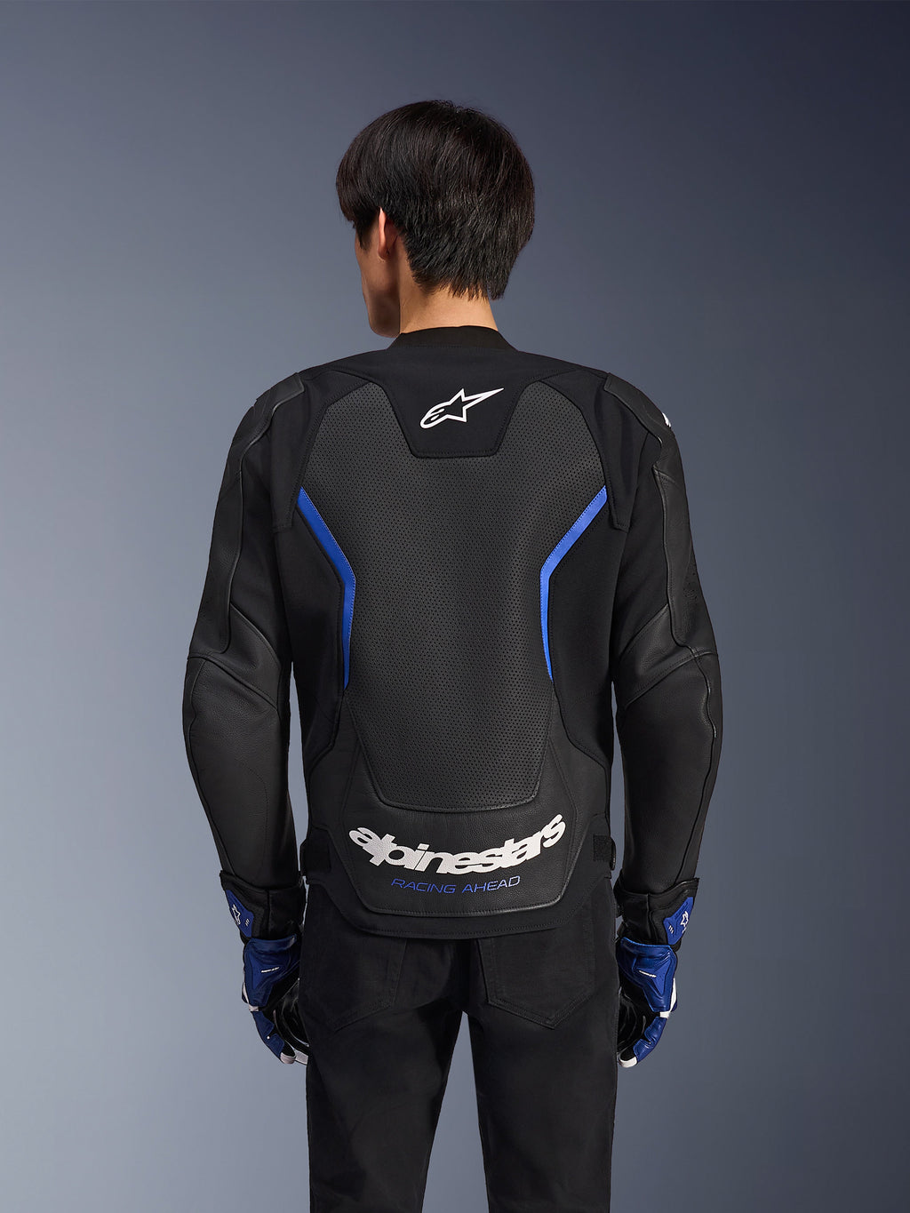 Alpinestars GP Force V2 Airflow Leather Jacket, skórzana kurtka motocyklowa, czarna z metalicznymi niebieskimi akcentami, widok z tyłu, duży perforowany panel na plecach dla wentylacji, białe logo Alpinestars, noszona z niebieskimi rękawicami i czarnymi spodniami do jazdy sportowej