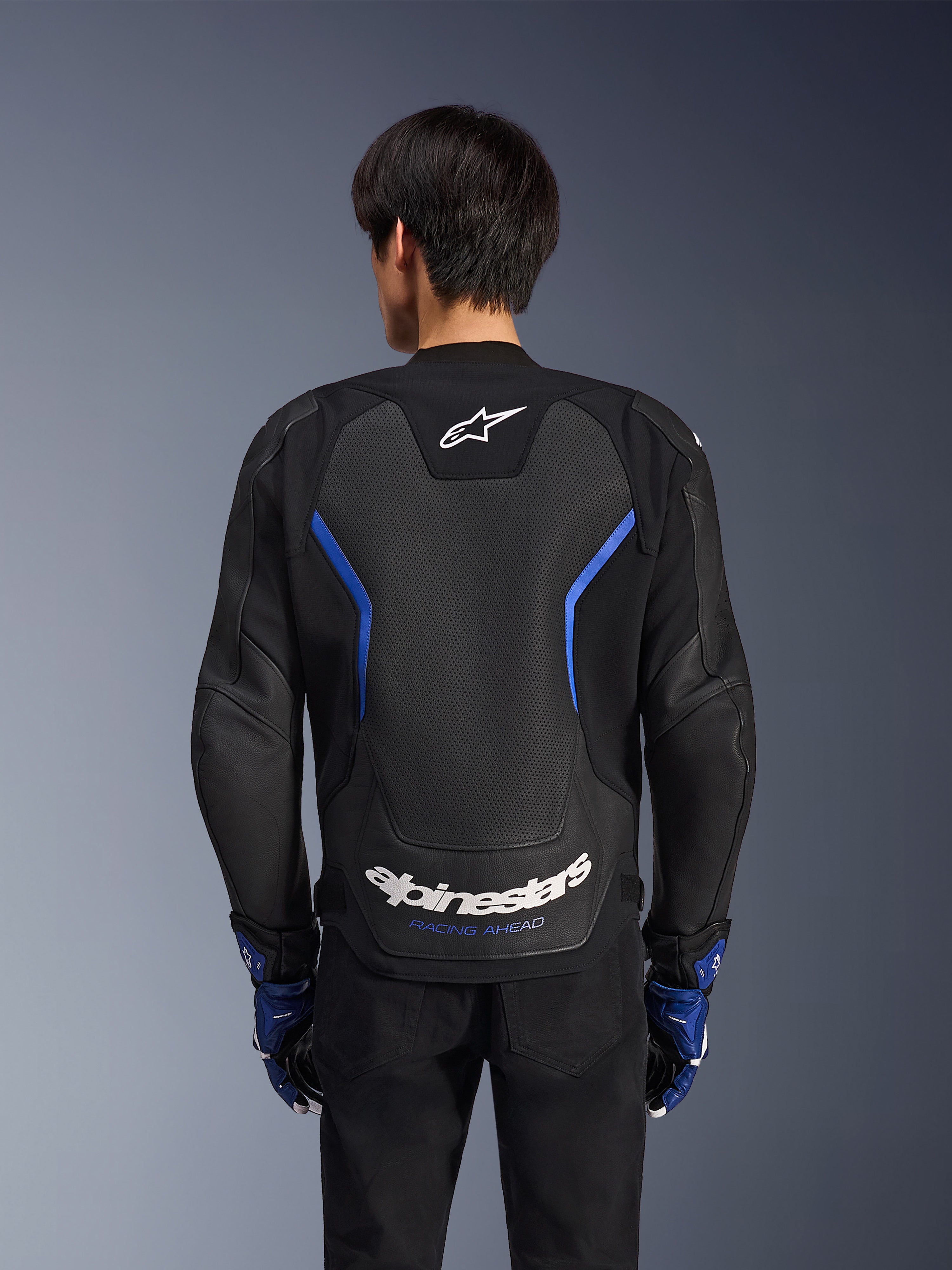 Alpinestars GP Force V2 Airflow Leather Jacket, skórzana kurtka motocyklowa, czarna z metalicznymi niebieskimi akcentami, widok z tyłu, duży perforowany panel na plecach dla wentylacji, białe logo Alpinestars, noszona z niebieskimi rękawicami i czarnymi spodniami do jazdy sportowej