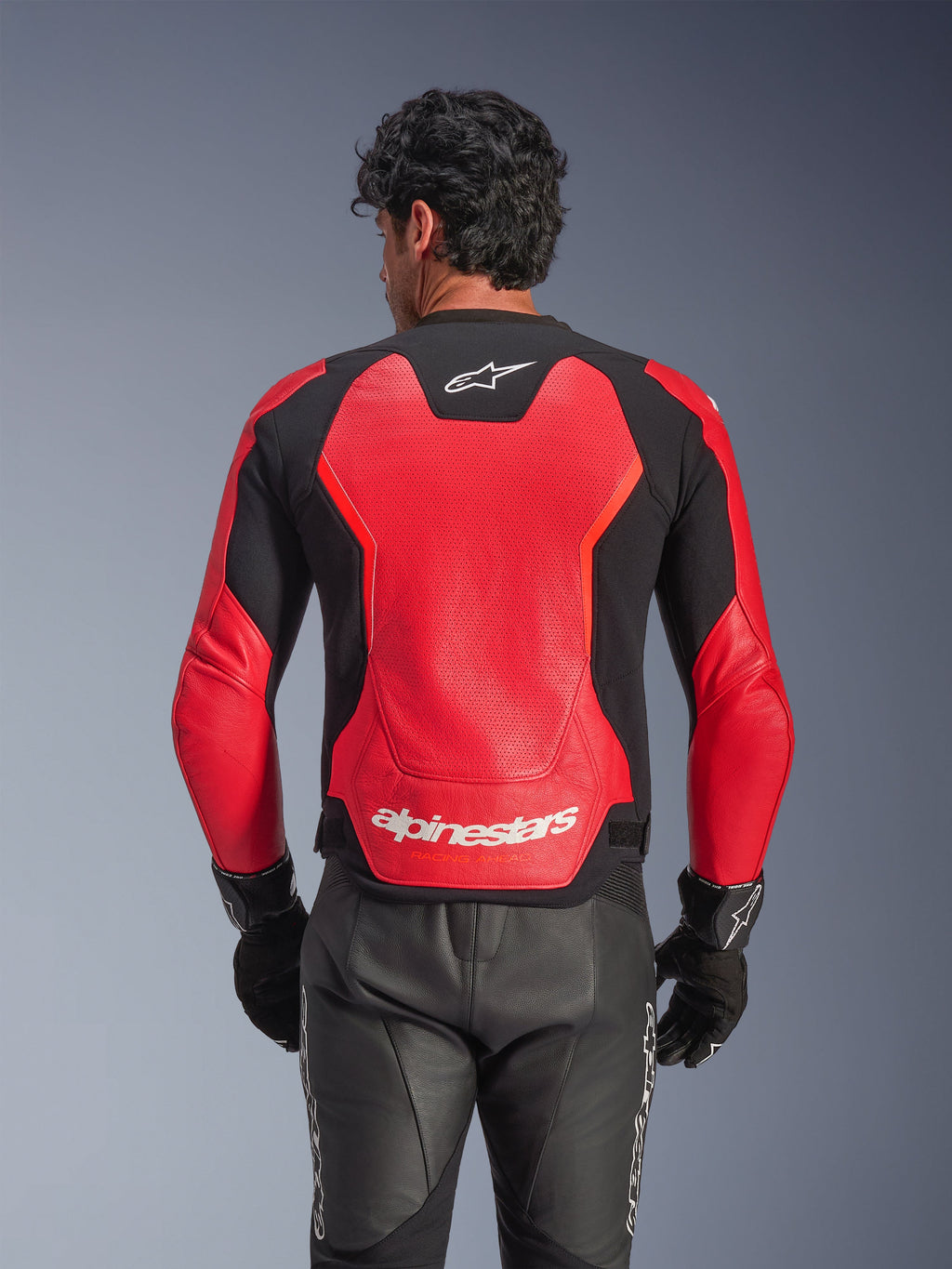 Kurtka skórzana Alpinestars GP Force V2 Air w kolorze czerwono-czarnym, widok z tyłu prezentujący perforowaną skórzaną konstrukcję zapewniającą przepływ powietrza, białe logo Alpinestars w dolnej części pleców oraz zintegrowane panele elastyczne, zestawiona z czarnymi skórzanymi spodniami motocyklowymi i rękawicami.