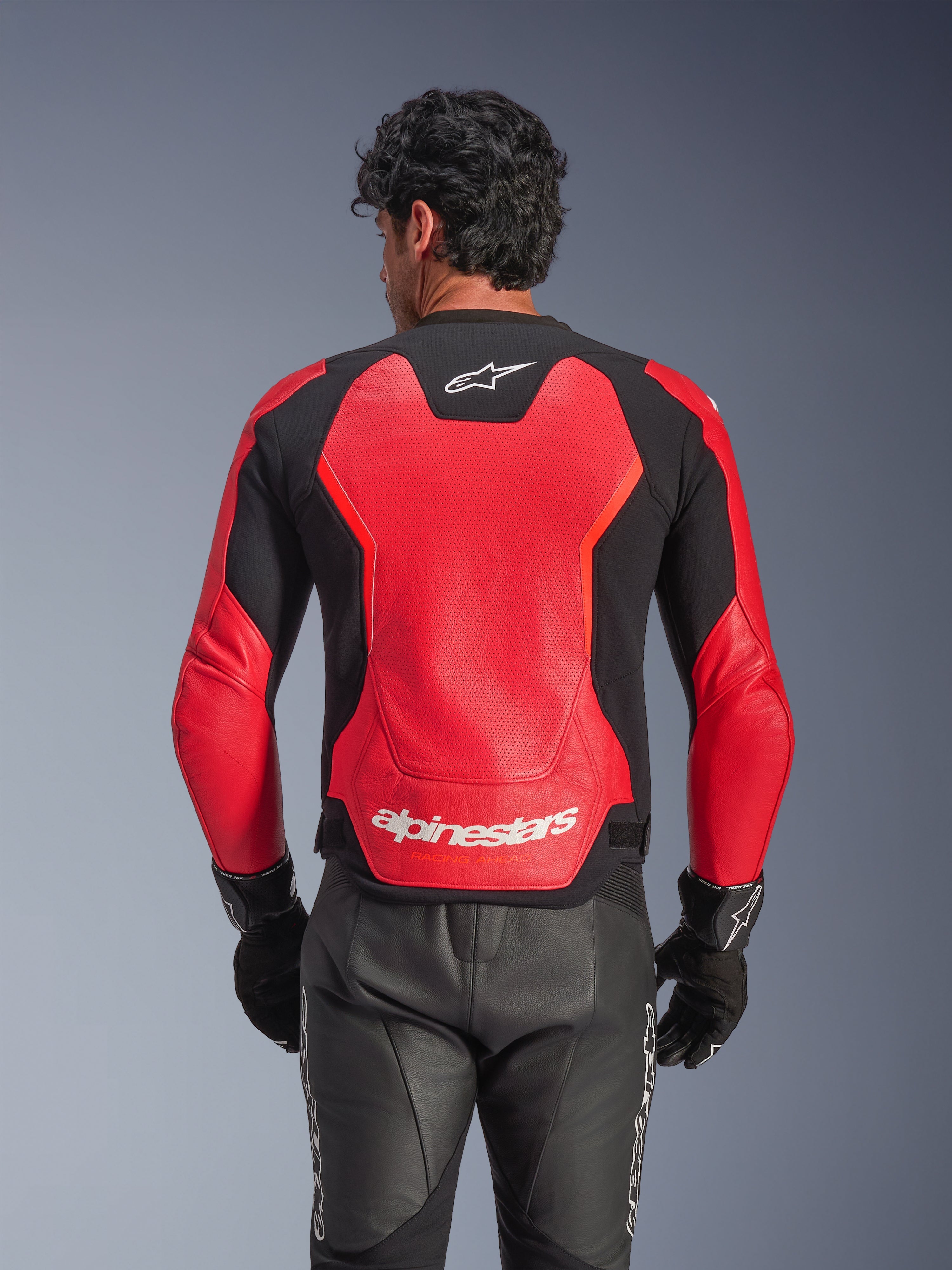 Kurtka skórzana Alpinestars GP Force V2 Air w kolorze czerwono-czarnym, widok z tyłu prezentujący perforowaną skórzaną konstrukcję zapewniającą przepływ powietrza, białe logo Alpinestars w dolnej części pleców oraz zintegrowane panele elastyczne, zestawiona z czarnymi skórzanymi spodniami motocyklowymi i rękawicami.