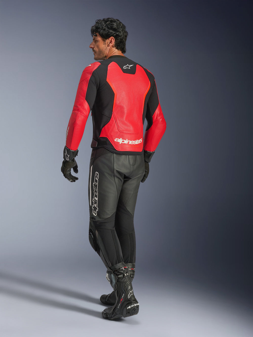Skórzana kurtka Alpinestars GP Force V2 Air, czerwono-czarna, widok z tyłu na motocyklistę, noszona z czarnymi skórzanymi spodniami i butami wyścigowymi, z perforowanymi panelami na plecach i brandingiem Alpinestars