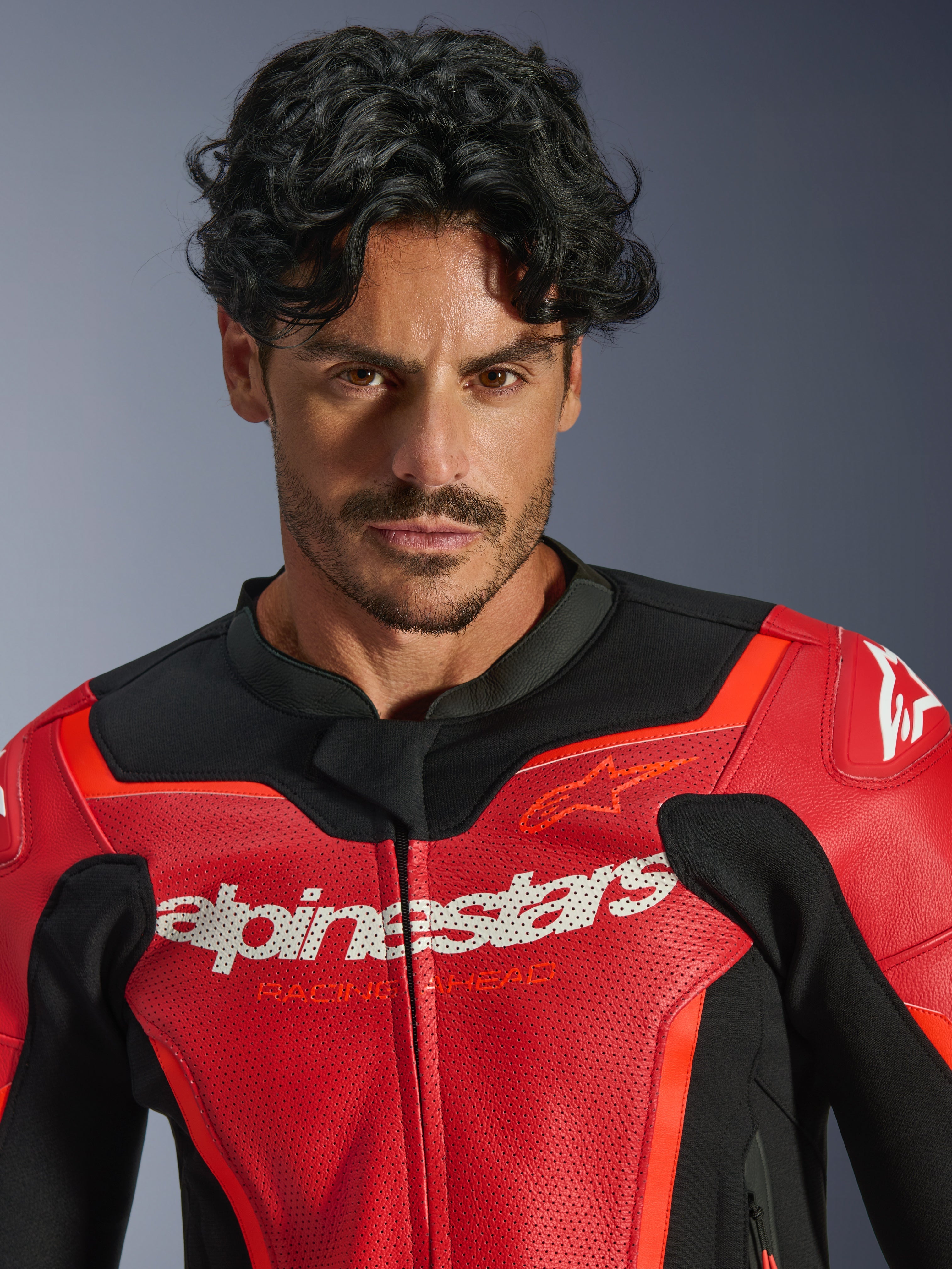 Skórzana kurtka Alpinestars GP Force V2 Air, czerwona i czerwona fluo, prezentowana na modelu, białe logo Alpinestars i napis „Racing Ahead” na klatce piersiowej, perforowana skóra zapewniająca przepływ powietrza, sportowa odzież motocyklowa