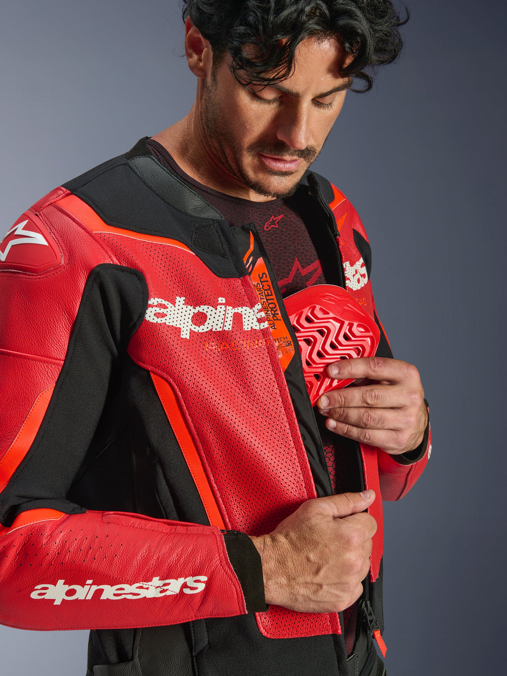 Kurtka skórzana Alpinestars GP Force V2 Air, czerwona i czerwona fluo, zbliżenie na motocyklistę wkładającego czerwony ochraniacz klatki piersiowej do wewnętrznej kieszeni, z perforowaną skórą i logo Tech-Air Ready, zaawansowana sportowa ochrona motocyklowa.