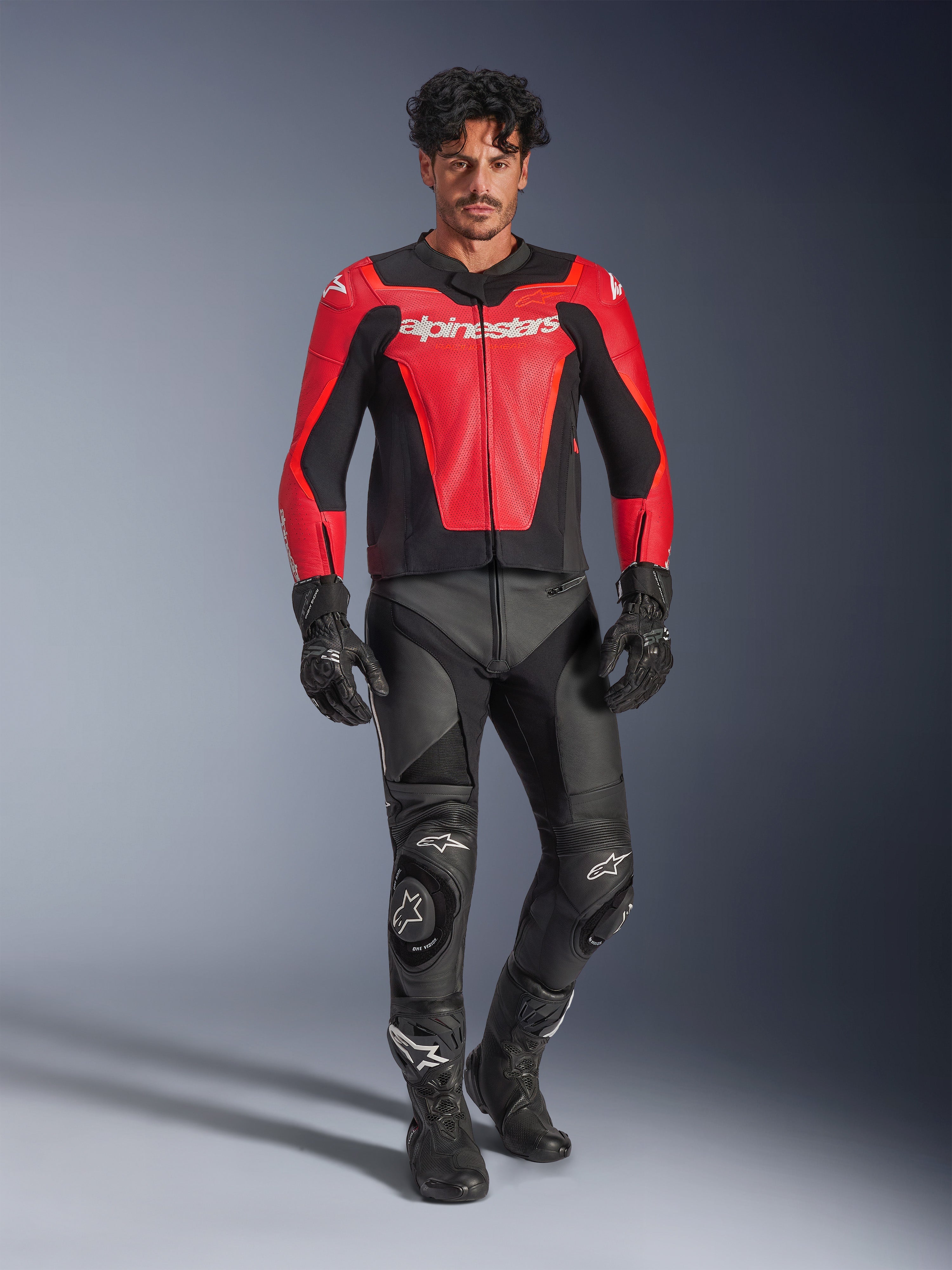 Kurtka skórzana Alpinestars GP Force V2 Air, czerwono-czarna, perforowana skóra z białym logo, noszona przez modela z czarnymi skórzanymi spodniami wyścigowymi i rękawicami, profesjonalny sprzęt motocyklowy
