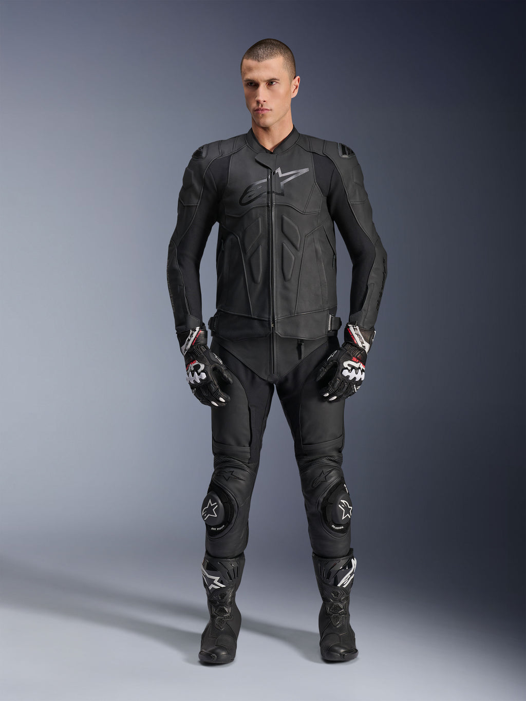 Alpinestars Dusk Leather Jacket, czarna skórzana kurtka, noszona przez modela w pełnym zestawie wyścigowym wraz ze skórzanymi spodniami, rękawicami i butami, ergonomiczny krój, ochraniacze ramion i logo na klatce piersiowej.