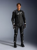 Męski model mający na sobie skórzaną kurtkę Alpinestars Dusk w kolorze czarno-białym, z wyraźnymi białymi logo na klatce piersiowej i ramionach, w zestawie z dopasowanymi czarnymi skórzanymi spodniami motocyklowymi, czarno-białymi rękawicami i czarnymi butami, kompletny sportowy strój wyścigowy do użytku drogowego.